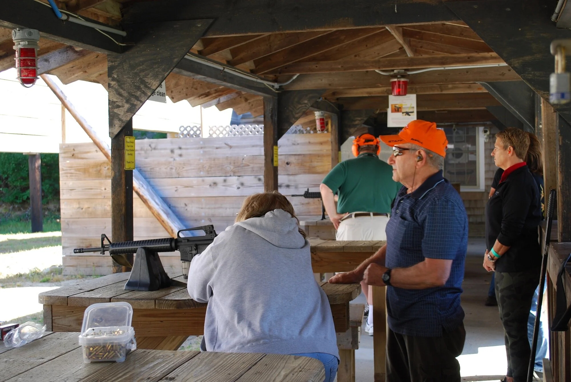 People at an outdoor shooting range, with one woman and a man in a blue shirt and orange hat observing, and others in the background preparing to shoot or watching. Firearms and ammunition are visible on the table.