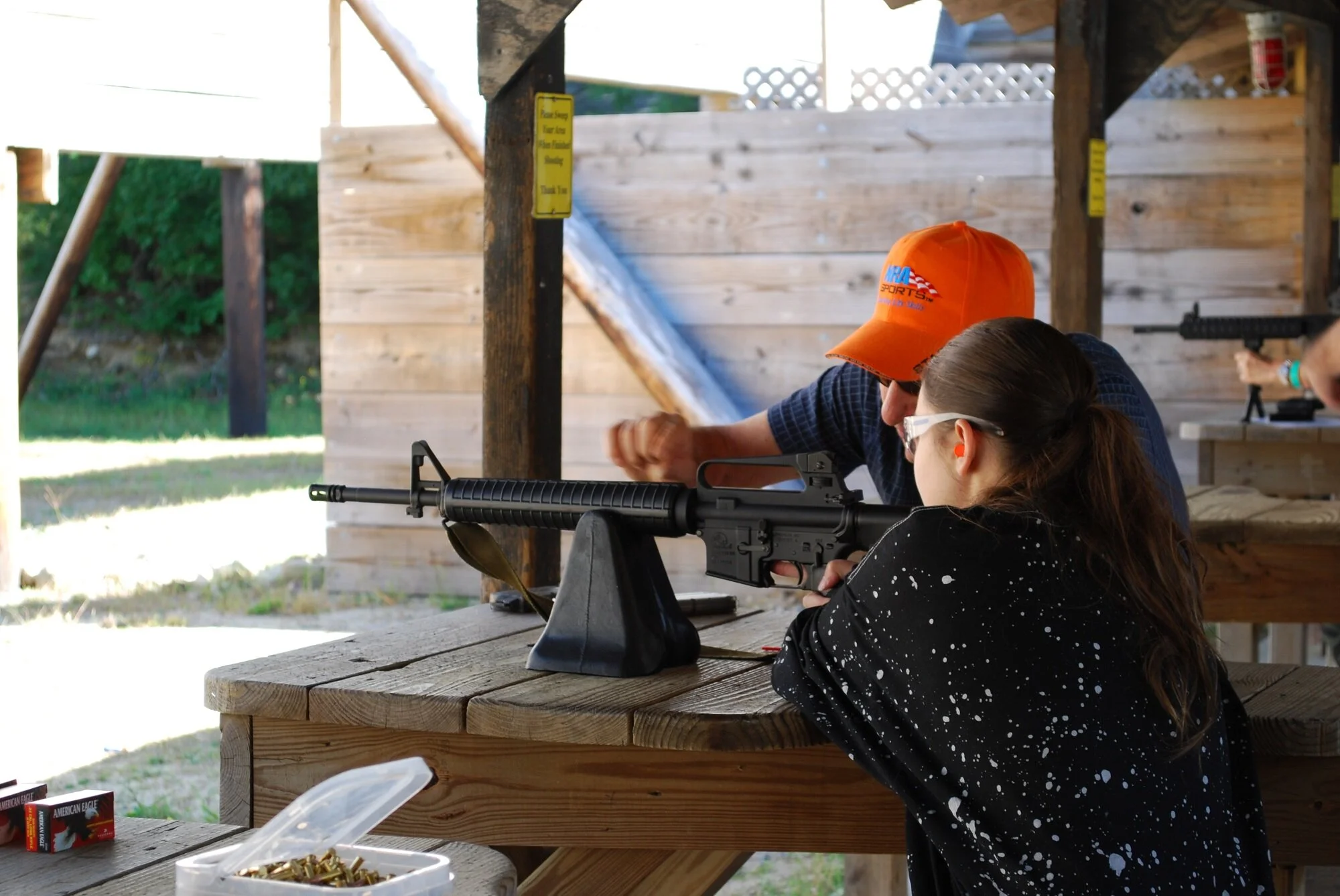 Child girl with glasses aiming a rifle at a shooting range under an outdoor shelter, with an instructor or adult guiding behind her.