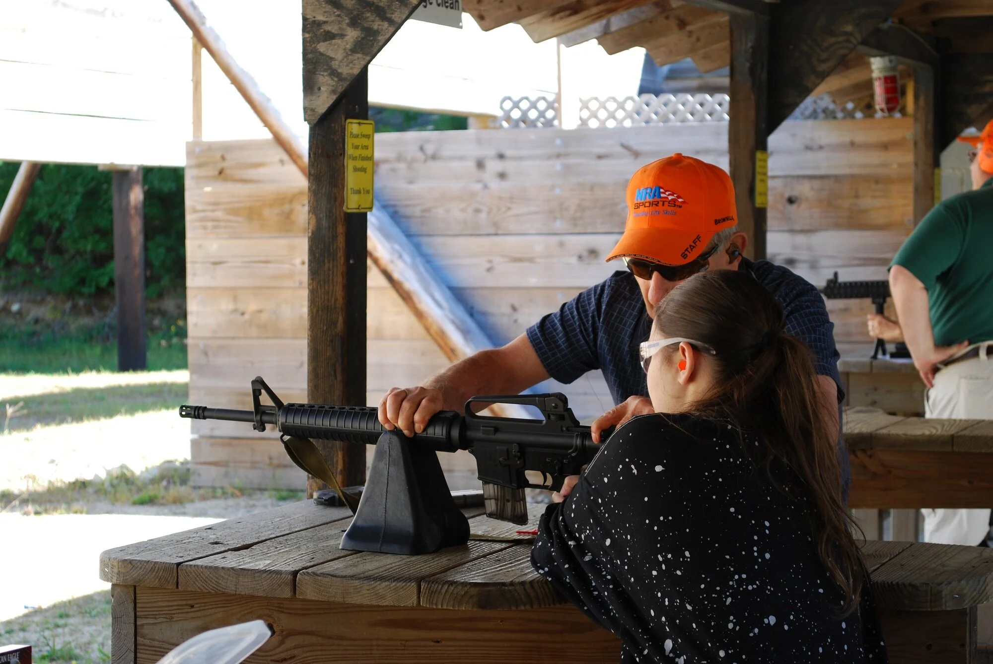 A woman and a man are at an outdoor shooting range. The woman is aiming a rifle while the man, wearing an orange hat and sunglasses, assists her. The background shows a wooden shelter with a sign and other individuals, including a person with a firea