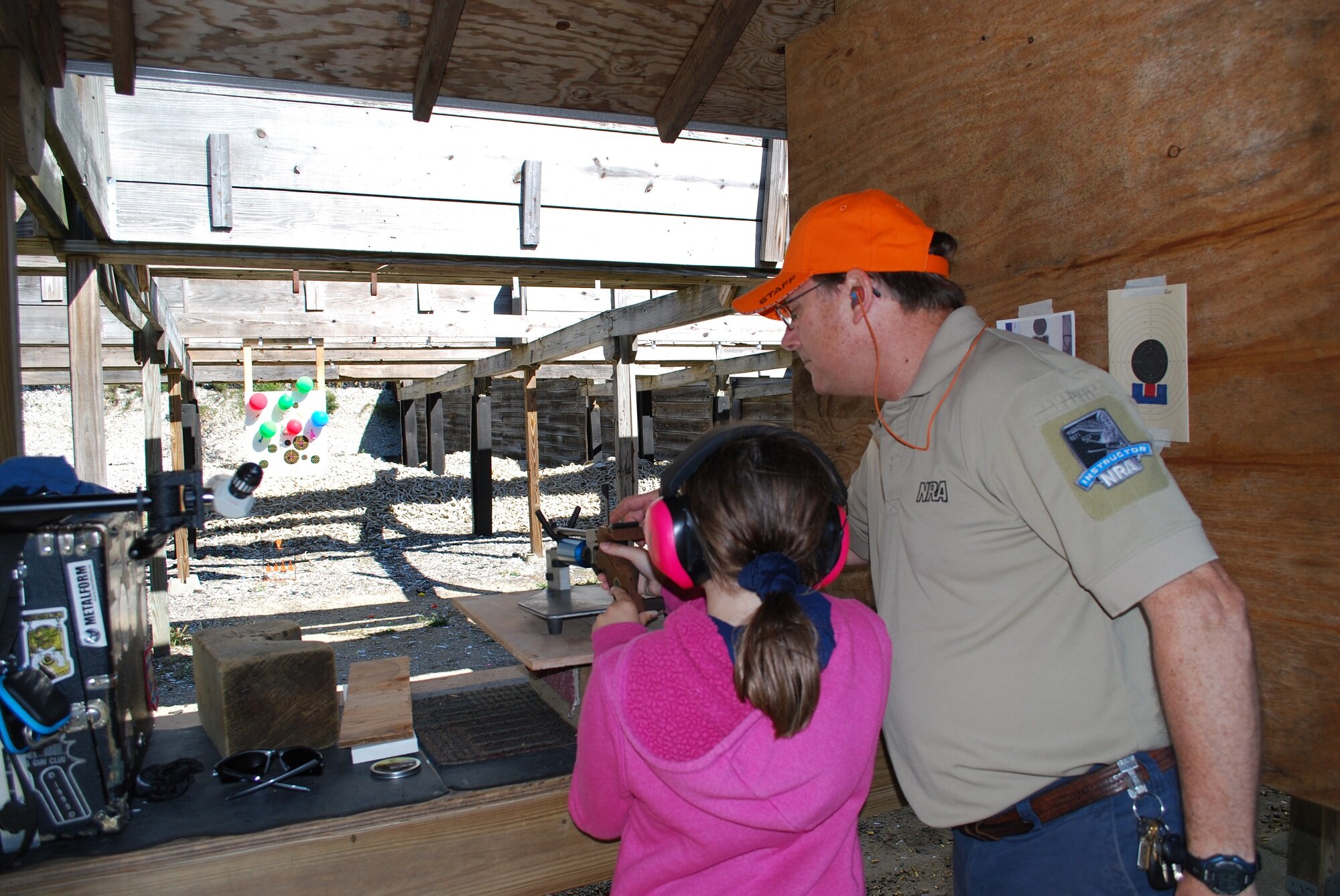 A young girl with pink earmuffs aiming a rifle at a shooting range under supervision of an instructor in an orange cap and beige shirt.
