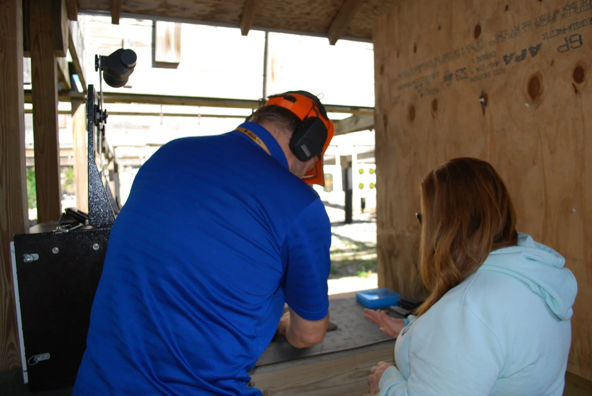 Two people at a shooting range, one man with orange safety headphones and a blue shirt leaning over a table and a woman with long red hair and white hoodie standing next to him, with wooden walls and shooting targets in the background.