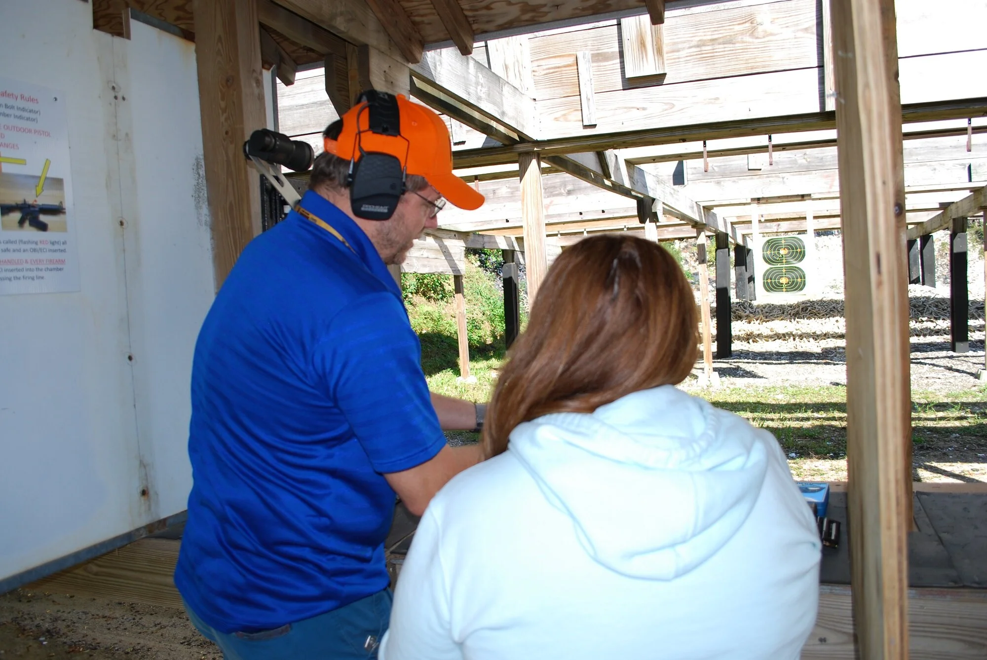 Man in blue shirt and orange cap and woman with brown hair at shooting range, aiming at targets while under wooden shelter.