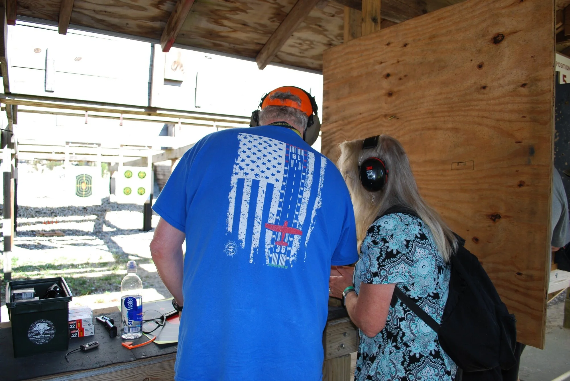 Two individuals wearing hearing protection at a shooting range, leaning over a table with various firearm accessories and supplies, with a target board in the background.