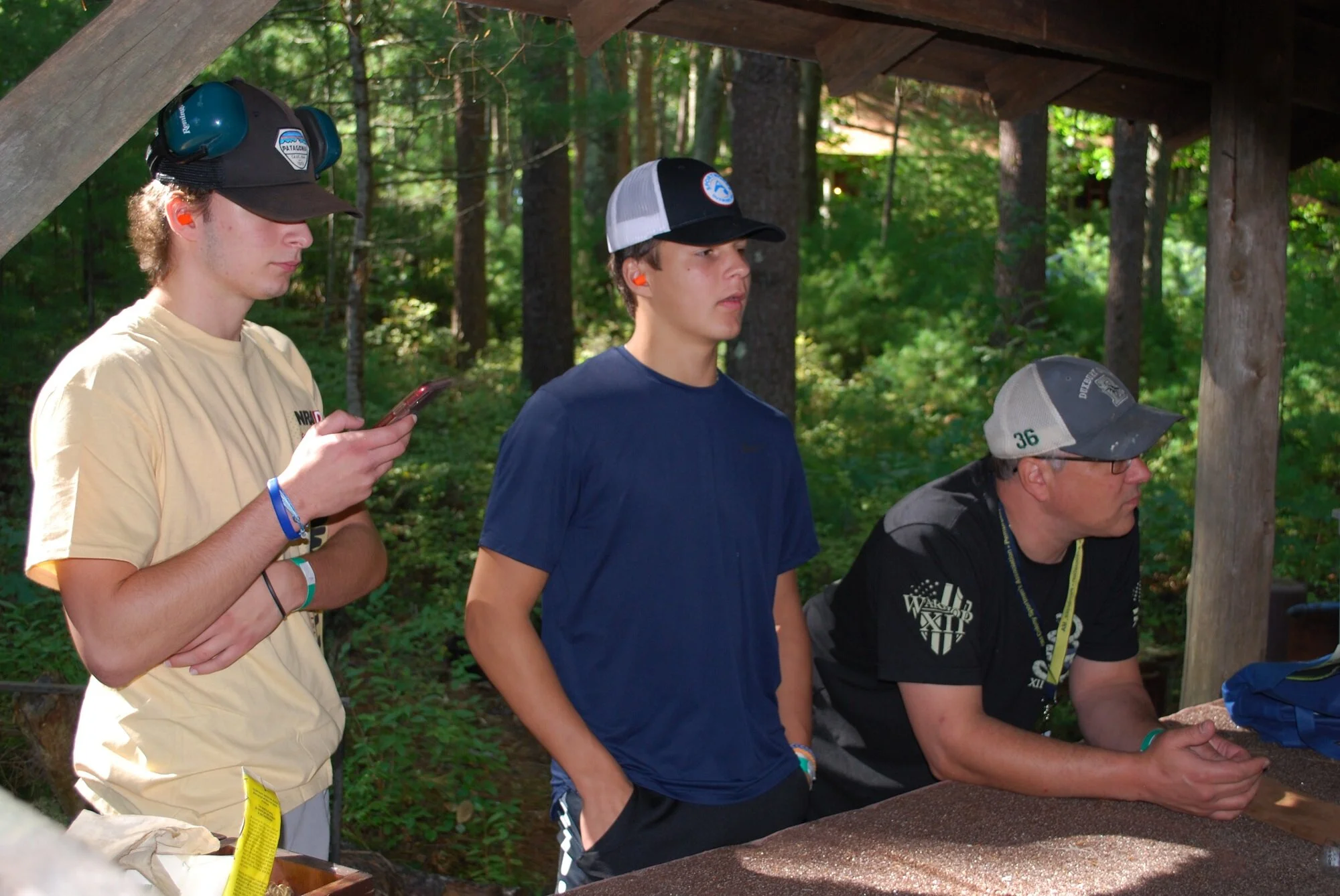 Three young men standing under a wooden shelter in a forest, with trees and green foliage in the background. One is looking at his phone, while the other two are looking forward or at each other.