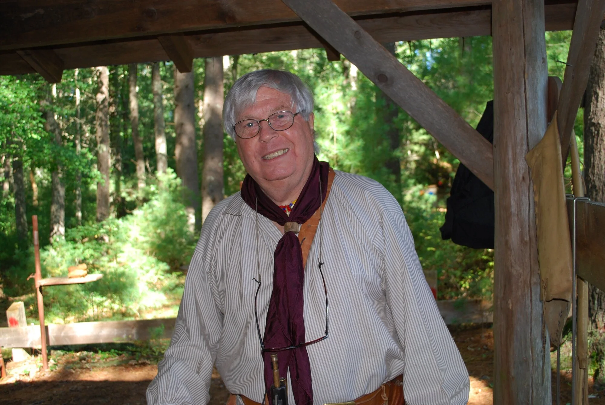 An older man with glasses and gray hair smiling outdoors under a wooden structure, surrounded by green trees.