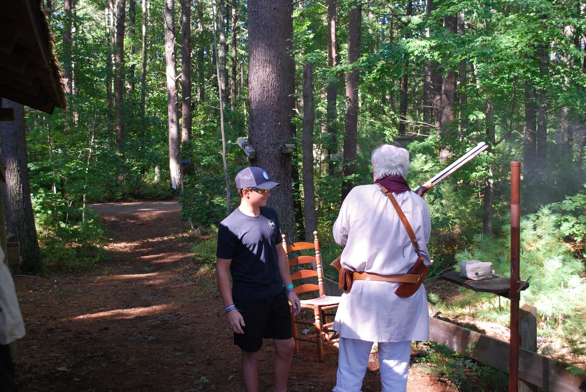 An elderly man with white hair and a gray suit is explaining something to a young boy in a hat and dark clothing in a wooded outdoor setting, with trees and a dirt path in the background.