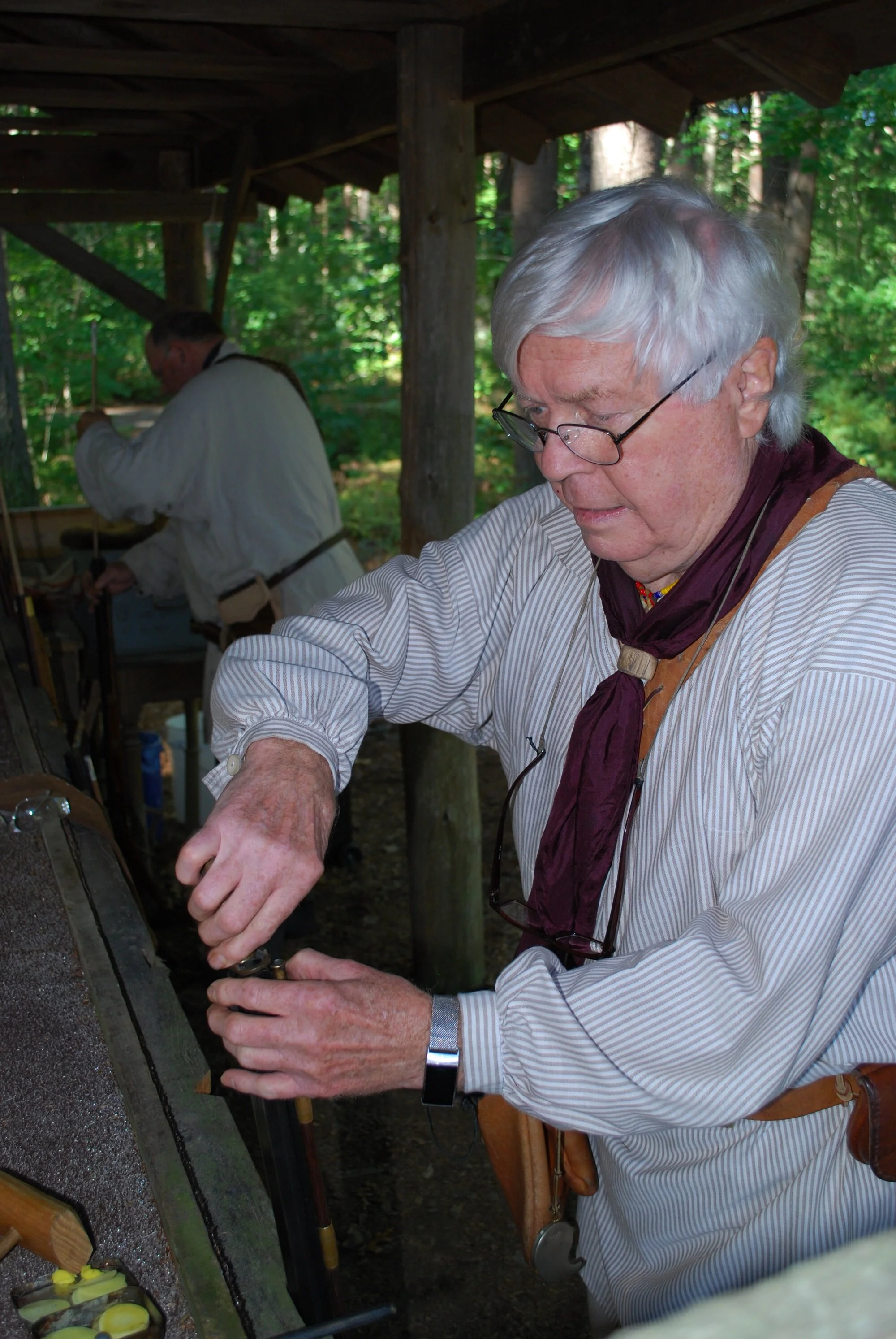 An elderly man with white hair, glasses, and a striped shirt is working with tools at an outdoor workspace under a wooden roof, with a person in historical attire in the background in a forest setting.