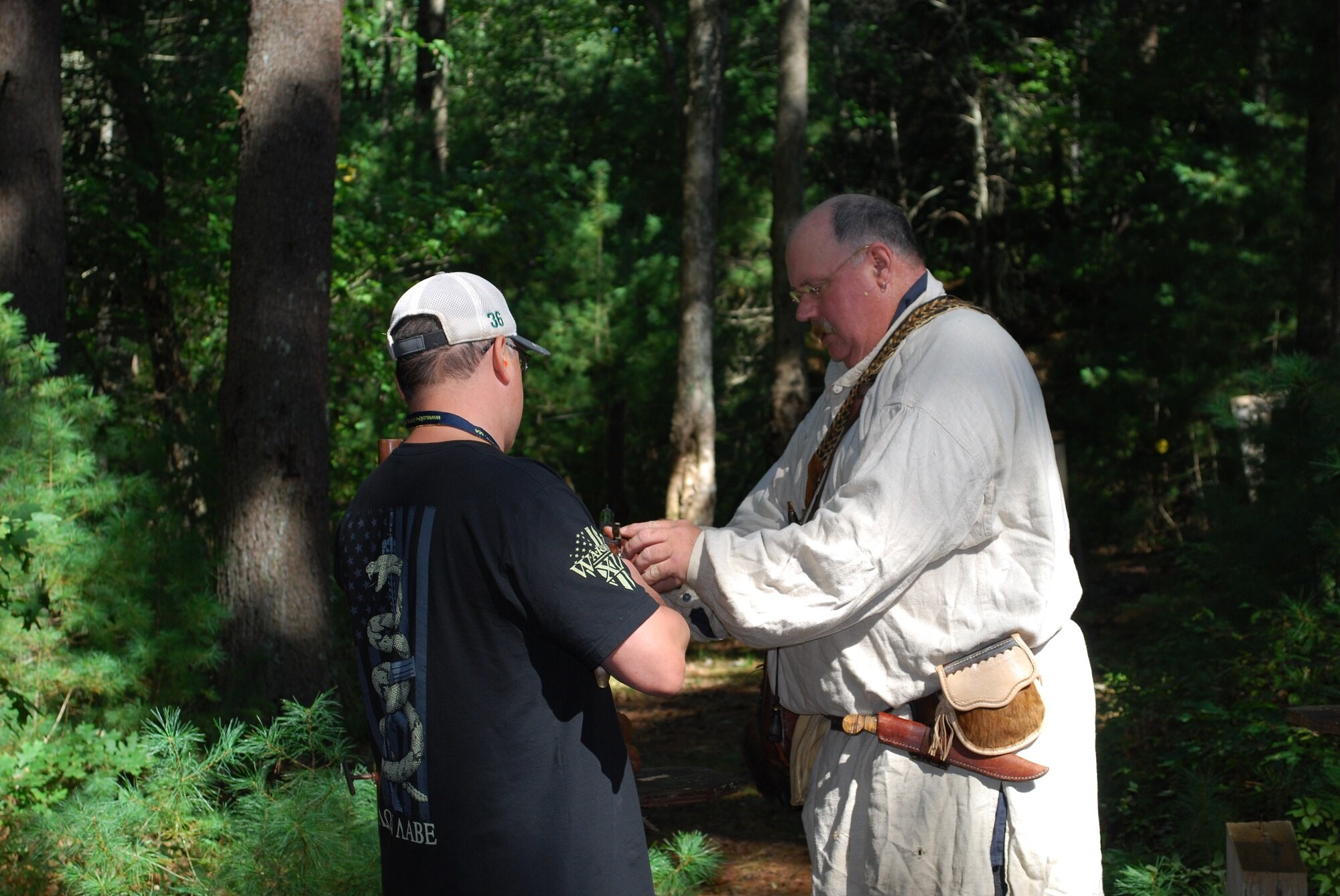 A young man and an older man in historical clothing standing in a forest, with the older man adjusting a strap or necklace around the young man's neck.