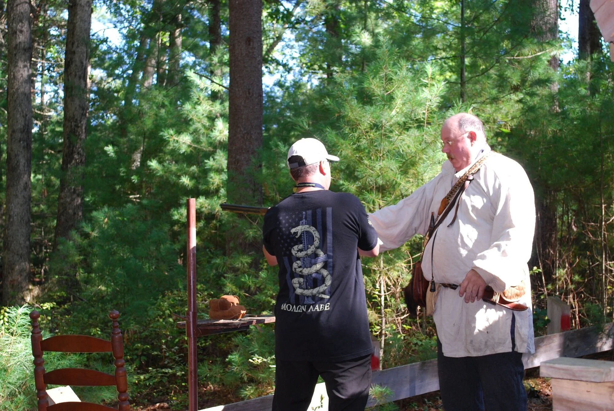 Two men in a forest, one dressed in a white period-style shirt and the other in a black t-shirt with a snake graphic, exchanging something.