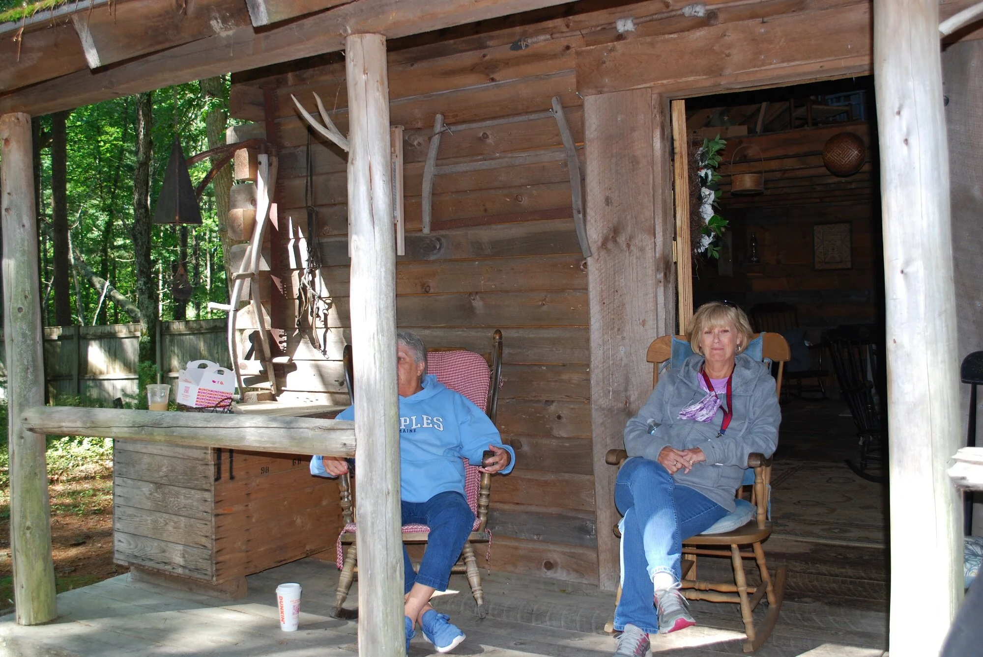 Two older women sitting outside on a porch in front of a rustic wooden cabin, surrounded by trees. One woman is wearing a gray hoodie and jeans, the other is in a blue hoodie and jeans. Various rustic tools hang on the cabin wall.