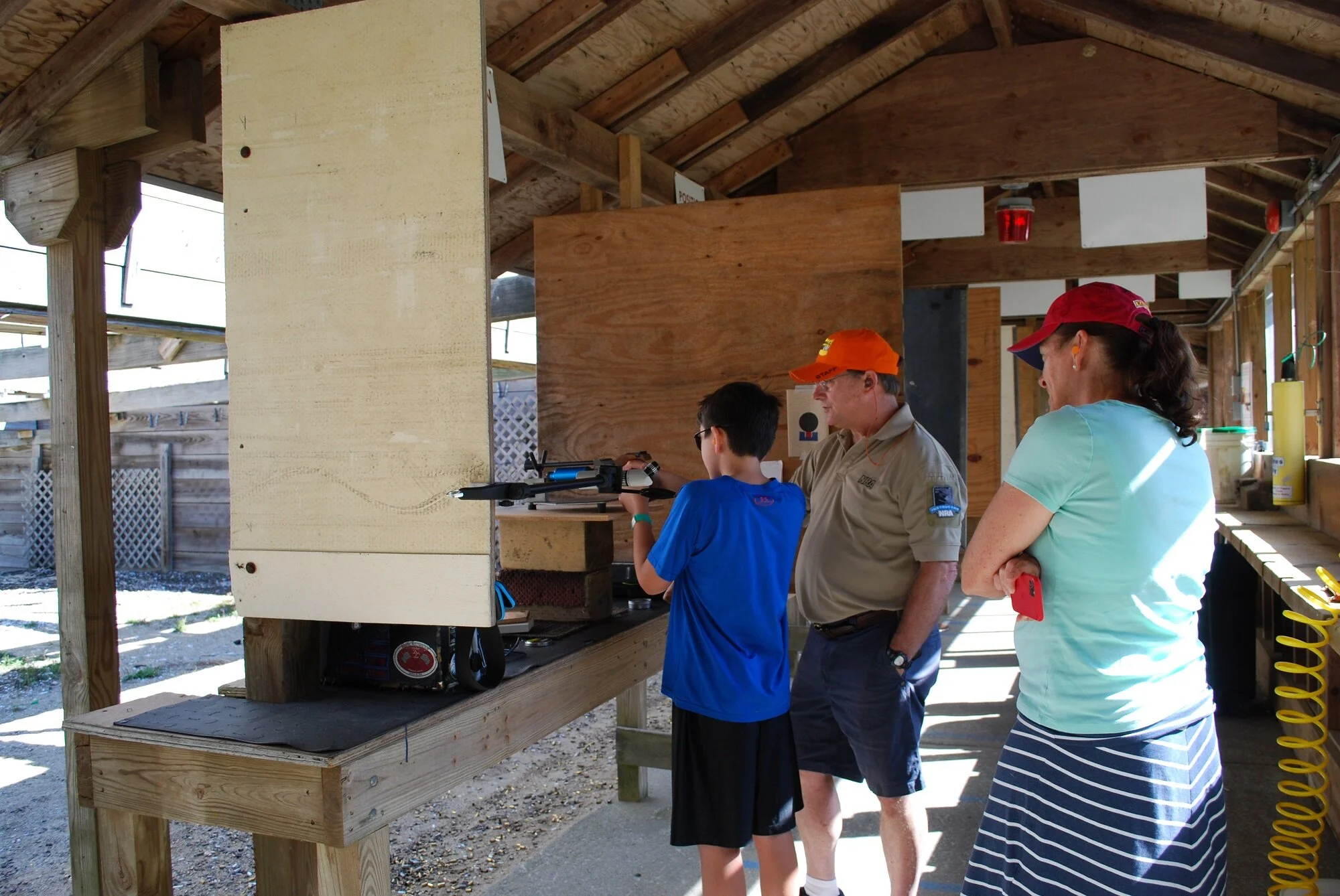 A young boy aiming a rifle at a shooting range supervised by an adult man, with a woman standing nearby, inside a wooden shelter.