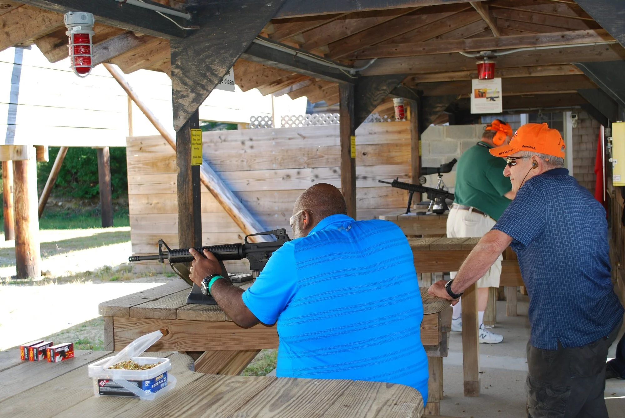 Three men at a shooting range practicing with rifles. One man in a blue shirt is aiming a rifle at a target, and two other men, wearing orange caps, are watching.