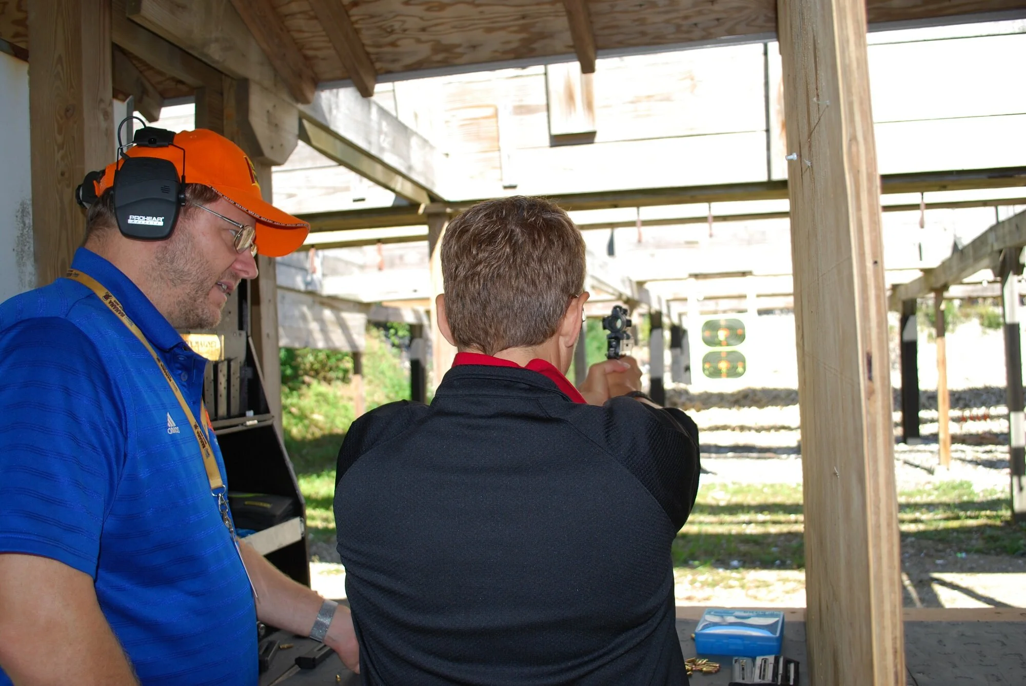 A man wearing an orange cap, blue shirt, and ear protection, is observing a person aiming a handgun at an outdoor shooting range with several target stands in the background.