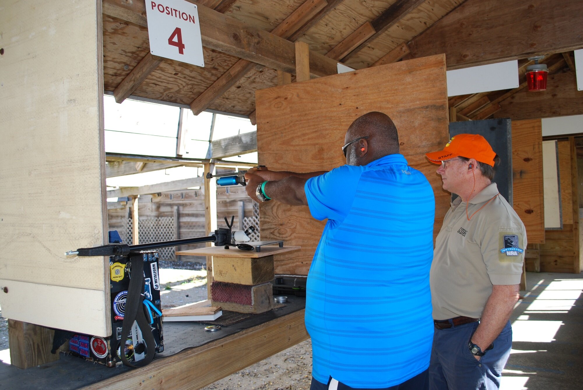 A man in a blue shirt aiming a rifle at a shooting range under a wooden shelter, with an instructor observing nearby. There is a sign that says 'Position 4' hanging above.