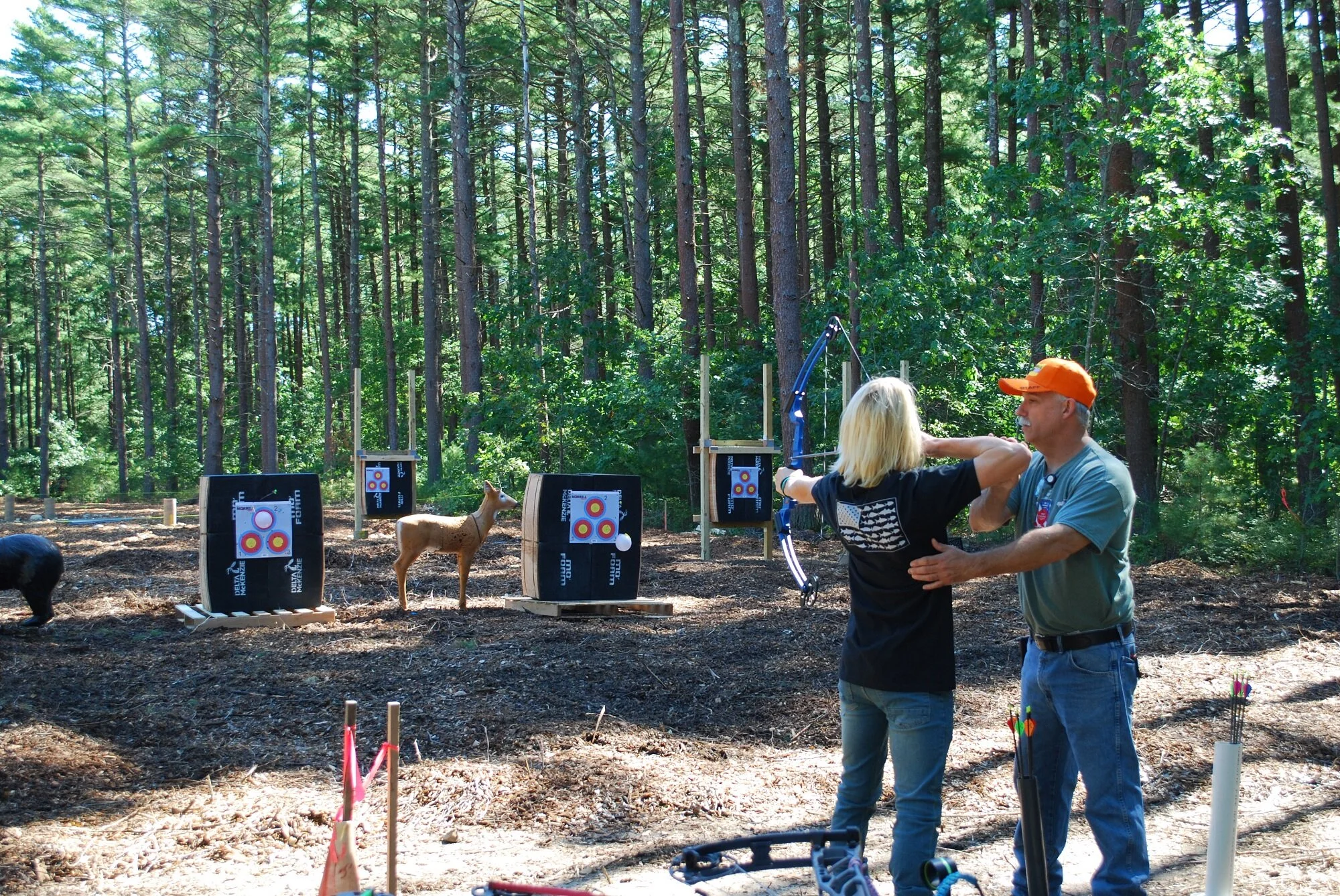 Two people practicing archery outdoors in a wooded area, with targets set up on stands.