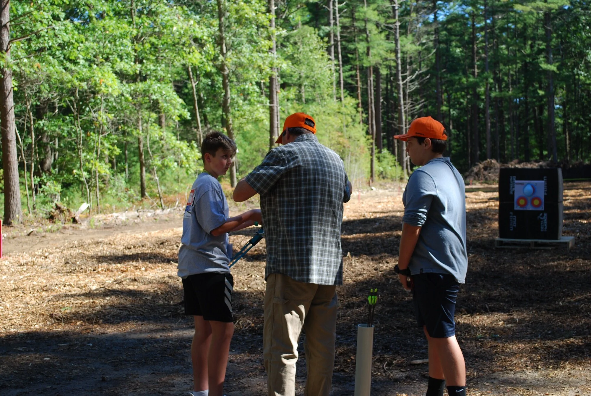 Three individuals, two boys and one adult man, are outdoors in a wooded area, engaged in an archery activity. The boy on the left is holding a bow and appears to be receiving instructions from the adult man in the middle, while the other boy on the r