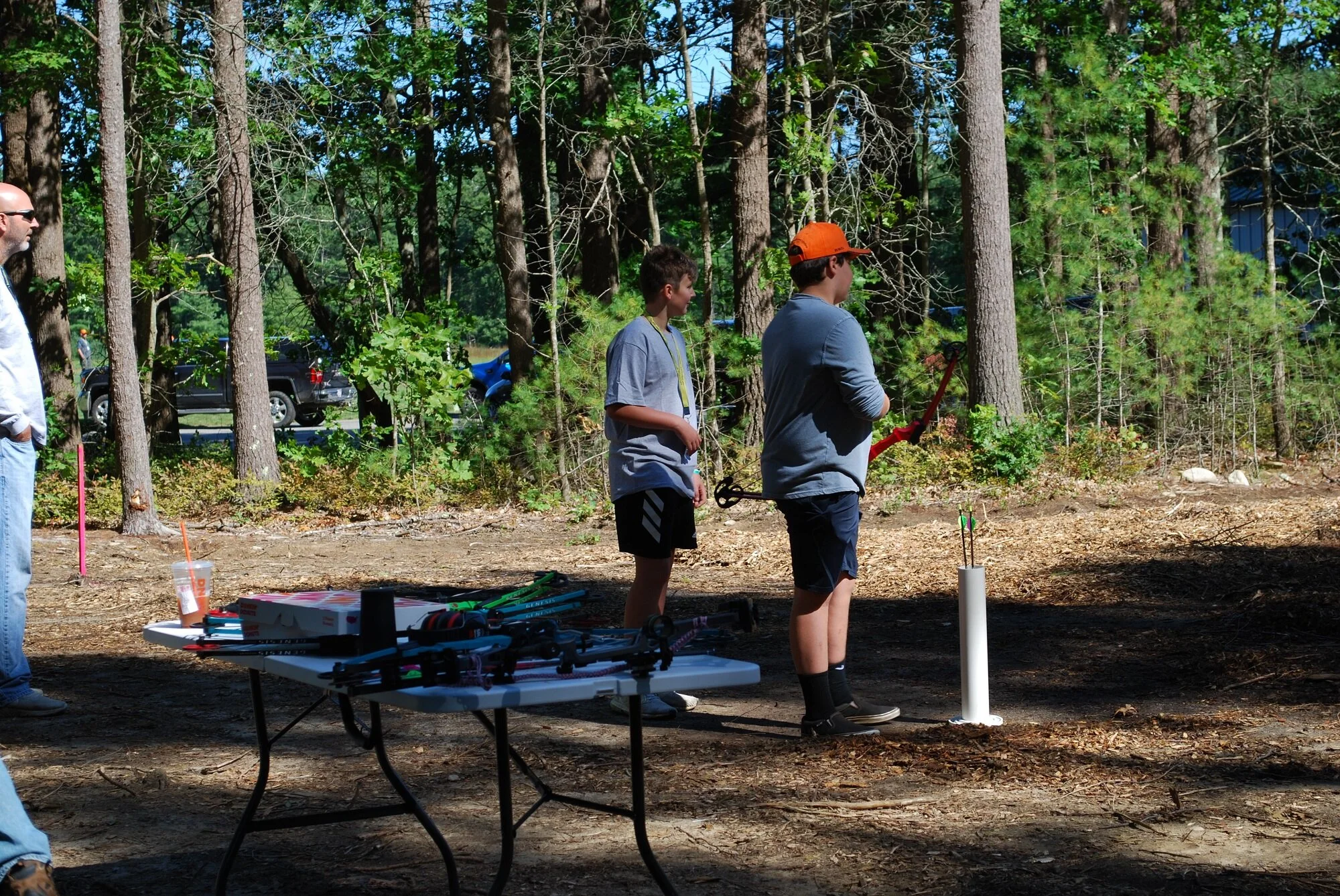 Two boys in a wooded outdoor area preparing to launch rockets, with a table of supplies nearby and an adult observing.