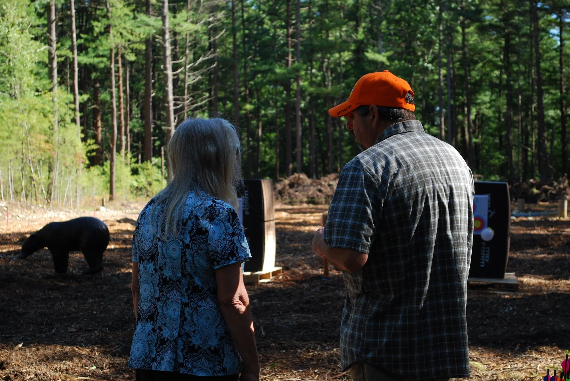 Two people, an older woman with long gray hair and a man wearing an orange cap and plaid shirt, standing outdoors in a wooded area, possibly discussing archery targets in the background. A black pig target is on the ground nearby.