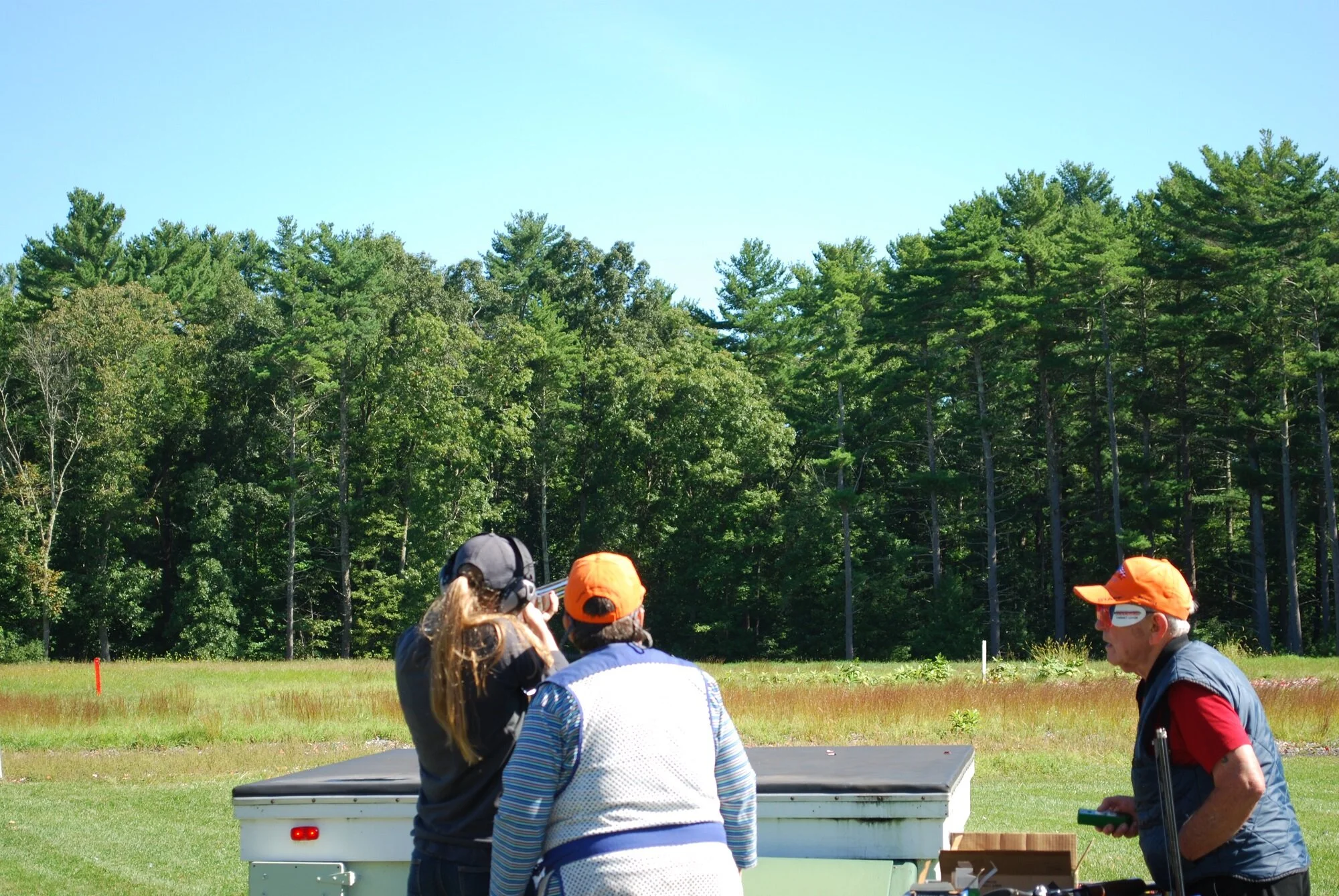 People at an outdoor shooting range with a wooded background, three individuals near a table, one holding a firearm, others observing, with a man on the right holding a control device.