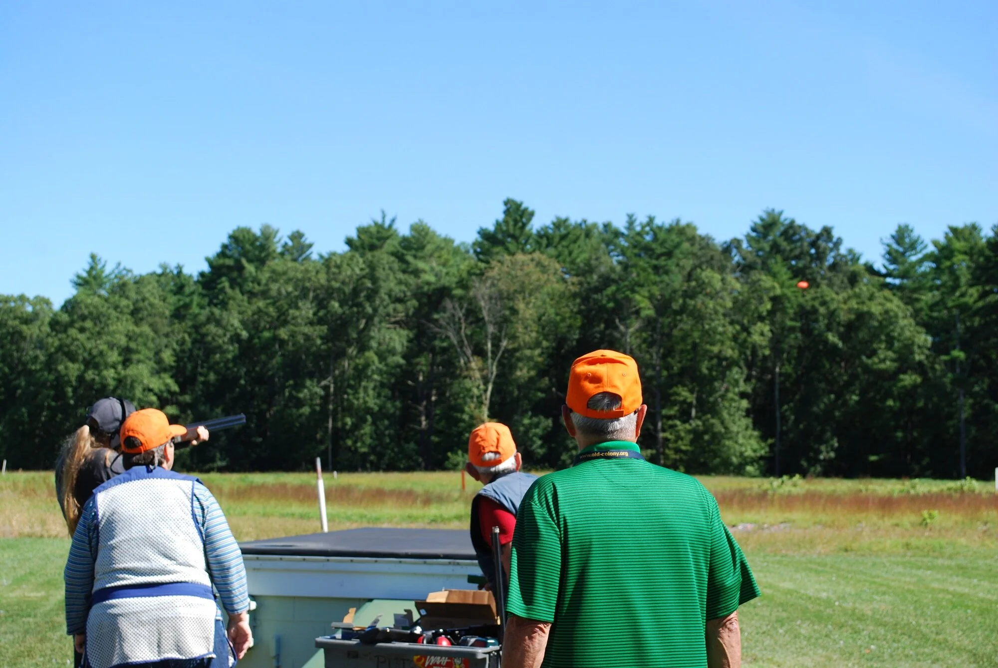 People outdoors at a shooting range, aiming shotguns, with a forest and blue sky in the background.