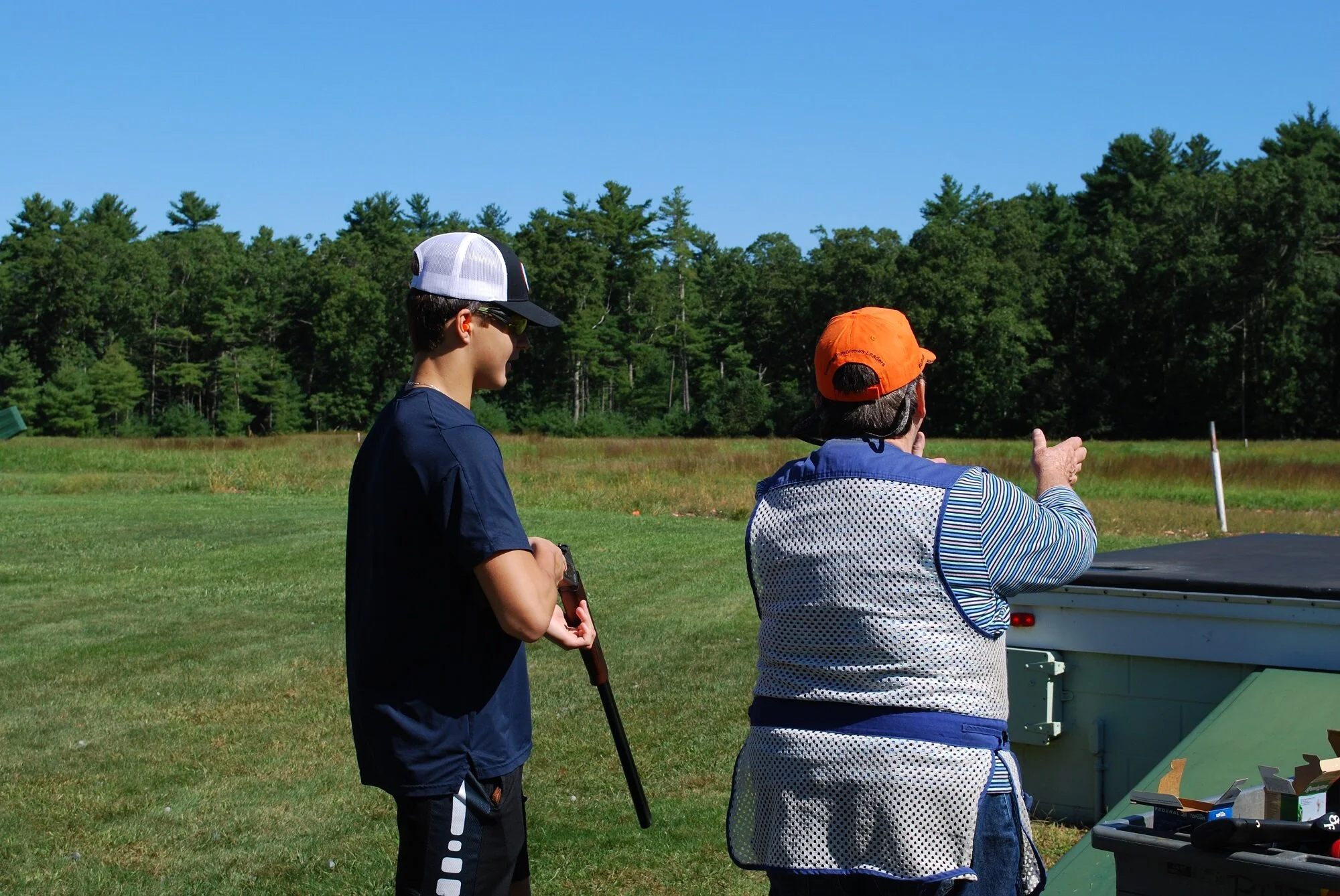 Two people, one young man and one older woman, at a shooting range outdoors. The young man is holding a shotgun, and the woman is pointing and guiding him. They are standing on a grassy field with trees in the background under a clear blue sky.