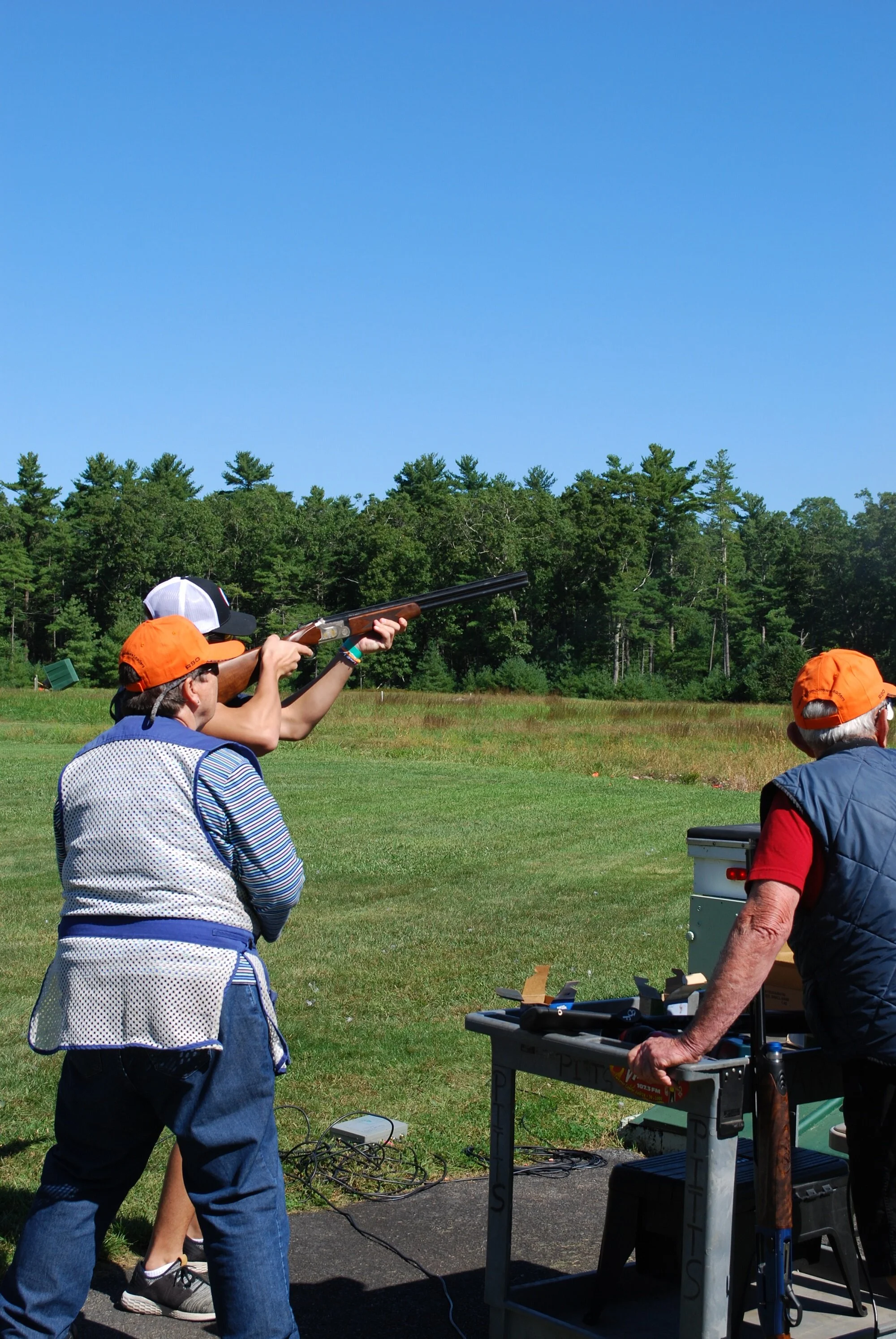Three people outdoors at a shooting range, with one person aiming a shotgun, and two others observing, amidst a green field and trees under a clear blue sky.