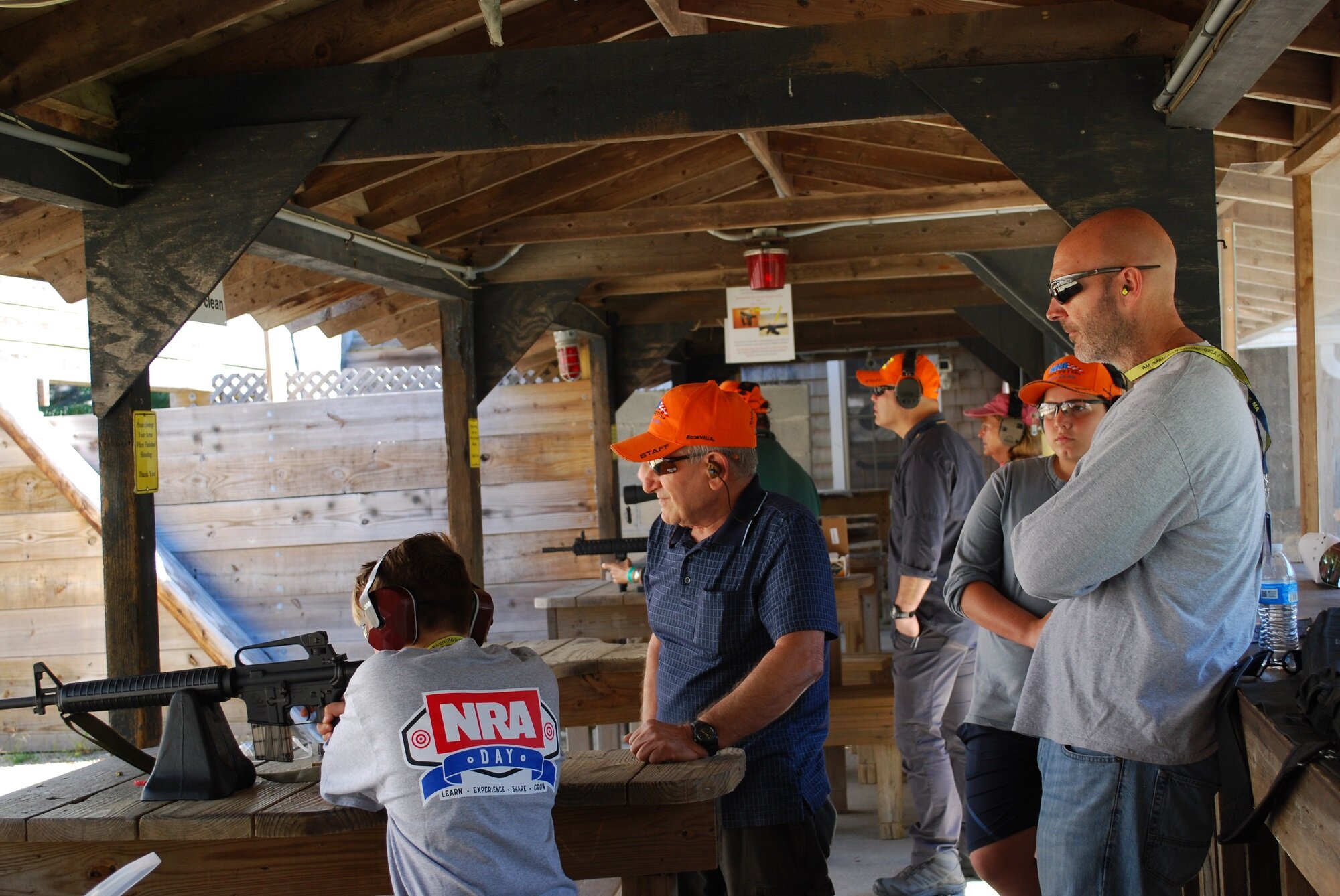 Group of people at an outdoor shooting range, with a boy aiming a rifle, others standing nearby, wearing protective ear and eye gear, and orange hats.