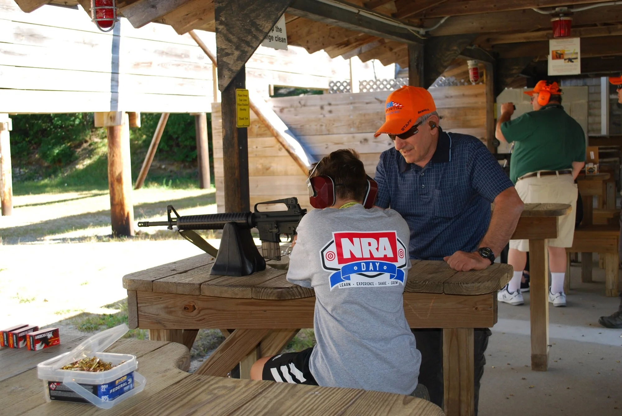 A young boy in a gray NRA Day t-shirt receiving shooting instructions from an older man at an outdoor shooting range. The boy is wearing ear protection and is seated at a wooden table with a rifle. The setting is covered with a roof, and other people