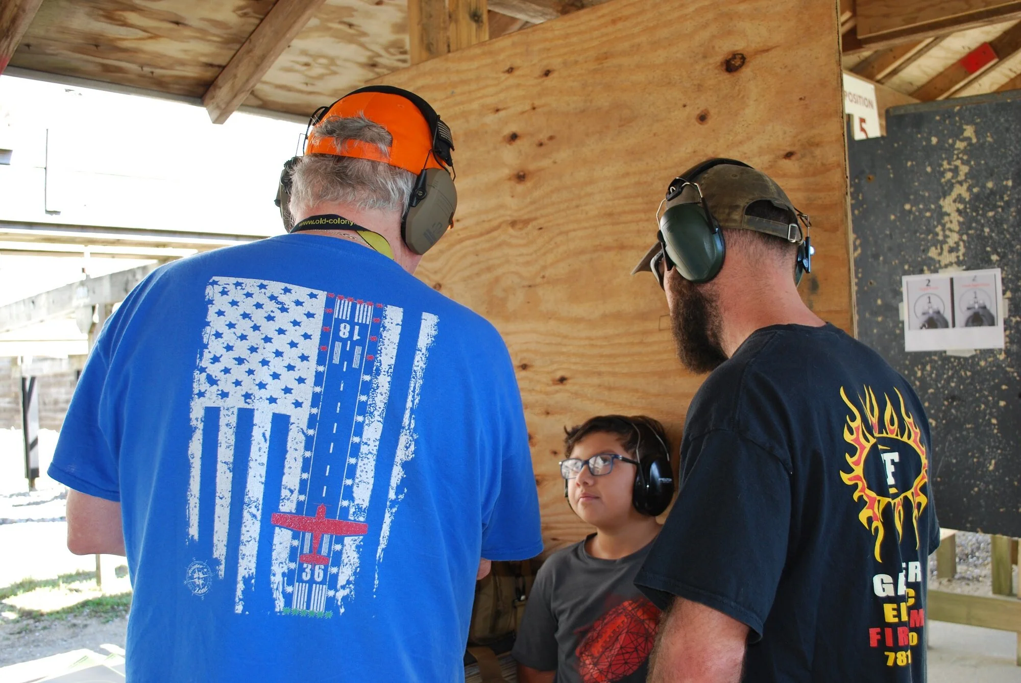 Two adult men and a boy at an outdoor shooting range, all wearing hearing protection. The man on the left has gray hair and is wearing an orange cap backwards and a blue T-shirt with an American flag and a red airplane on it. The man on the right has