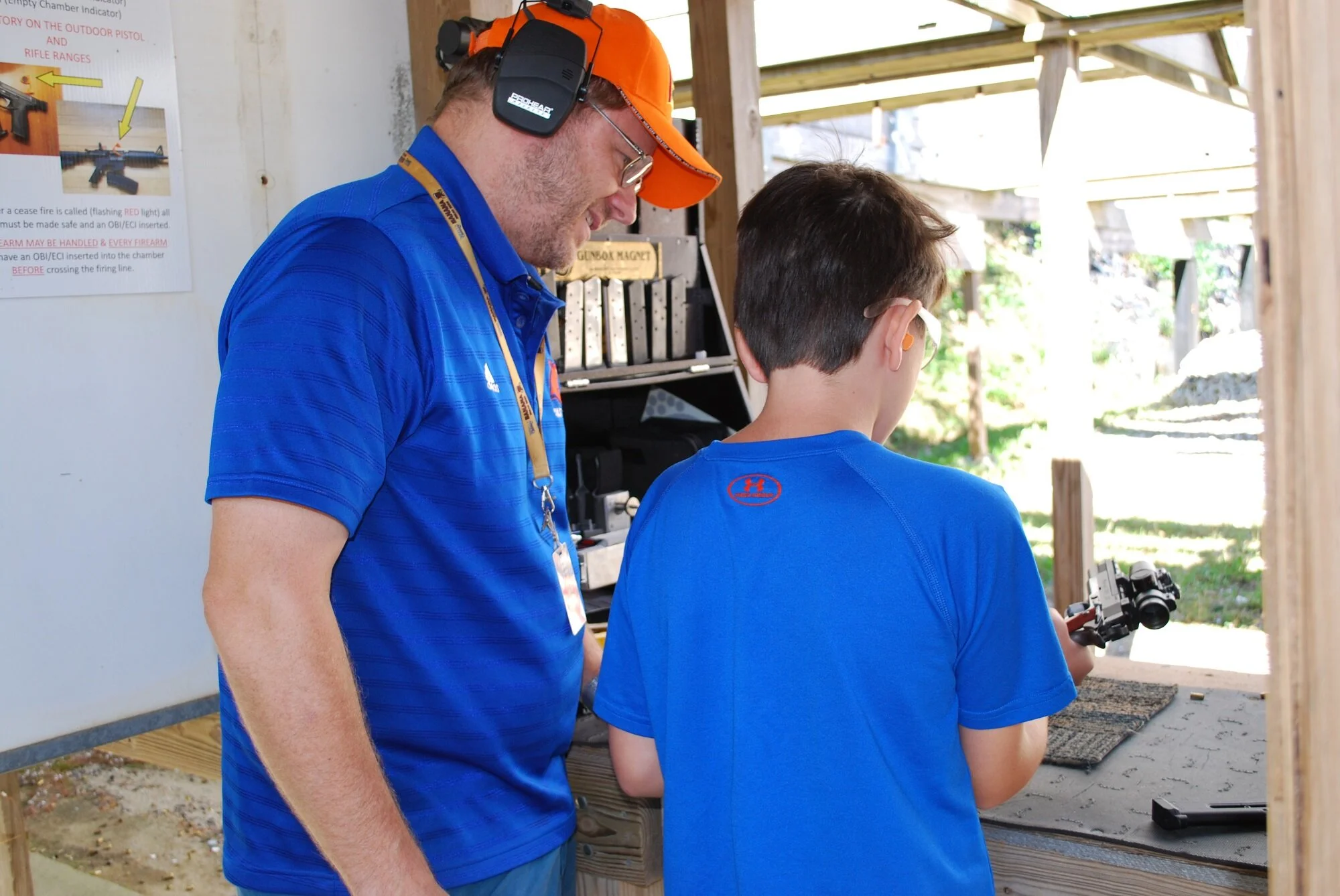 An instructor and a young boy at a shooting range. The instructor is wearing a blue shirt, orange cap, ear protection, and glasses, showing him instructing the boy. The boy is wearing a blue shirt, glasses, and ear protection, holding a handgun. They