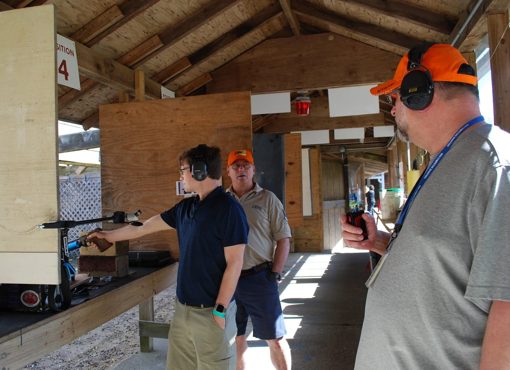 Three men at a shooting range with protective ear gear, one aiming a firearm, another observing, and the third speaking, under a wooden shelter with a sign labeled 'Position 4'.