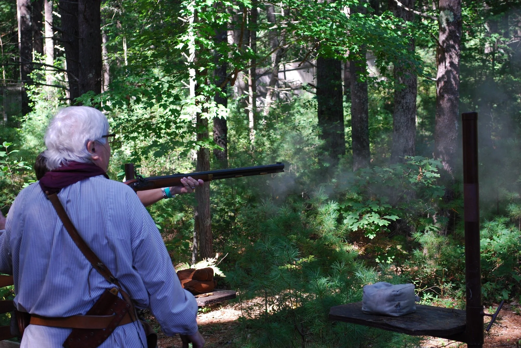 Older man shooting a rifle at a target in a forest during daytime.