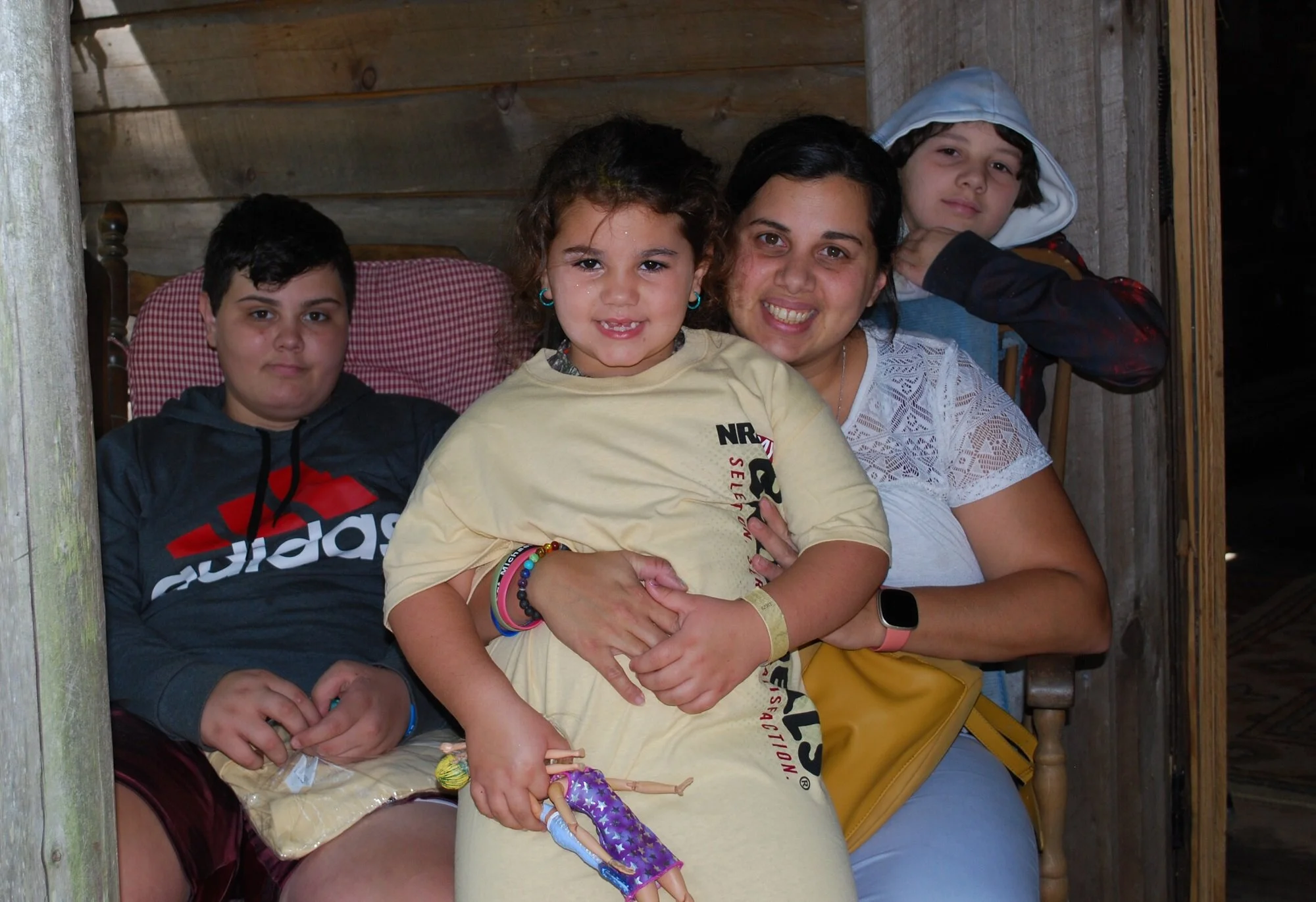 A woman and three children sitting on a wooden bench inside a rustic wooden structure. The woman and girl in front are smiling, while the two boys in the background have neutral expressions. The girl is holding a doll.