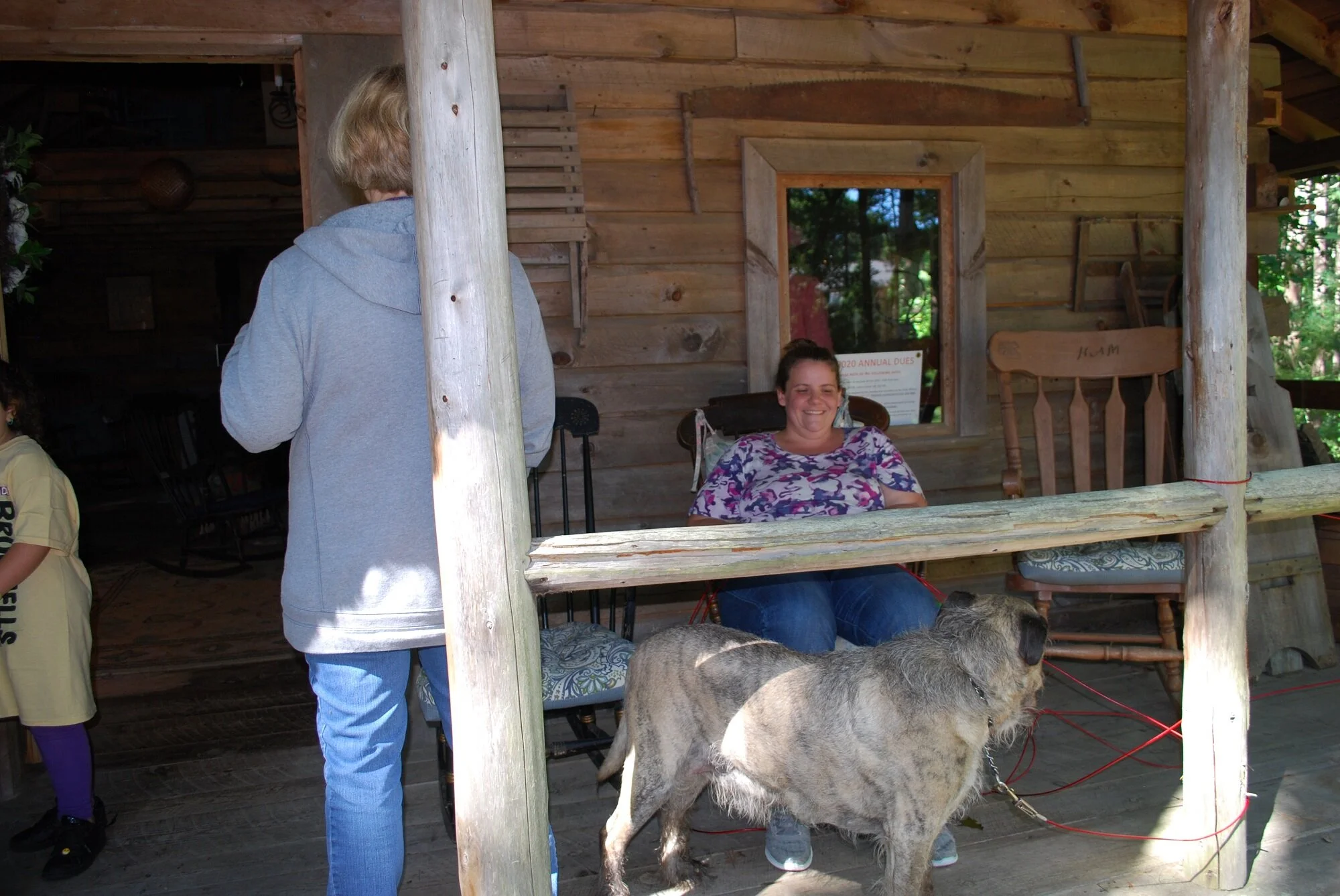 A woman sitting on a porch smiling, with a large dog tethered nearby. A man stands with his back to the camera, and part of a girl in a yellow shirt and purple leggings is visible to the left.