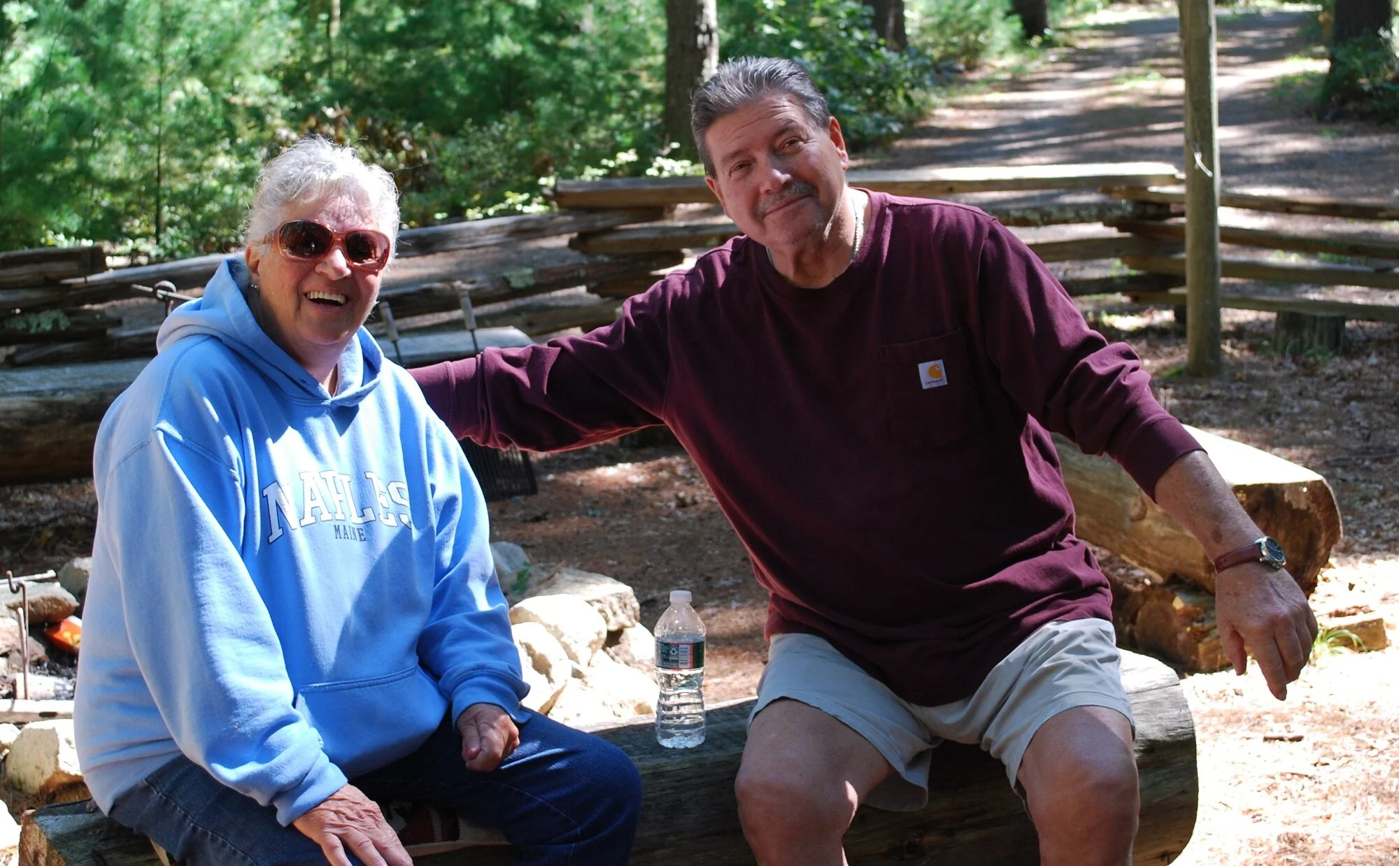 A smiling elderly woman wearing sunglasses and a blue hoodie with white text, and a middle-aged man in a maroon shirt and shorts, sitting outdoors on a wooden bench surrounded by trees, with a water bottle between them.