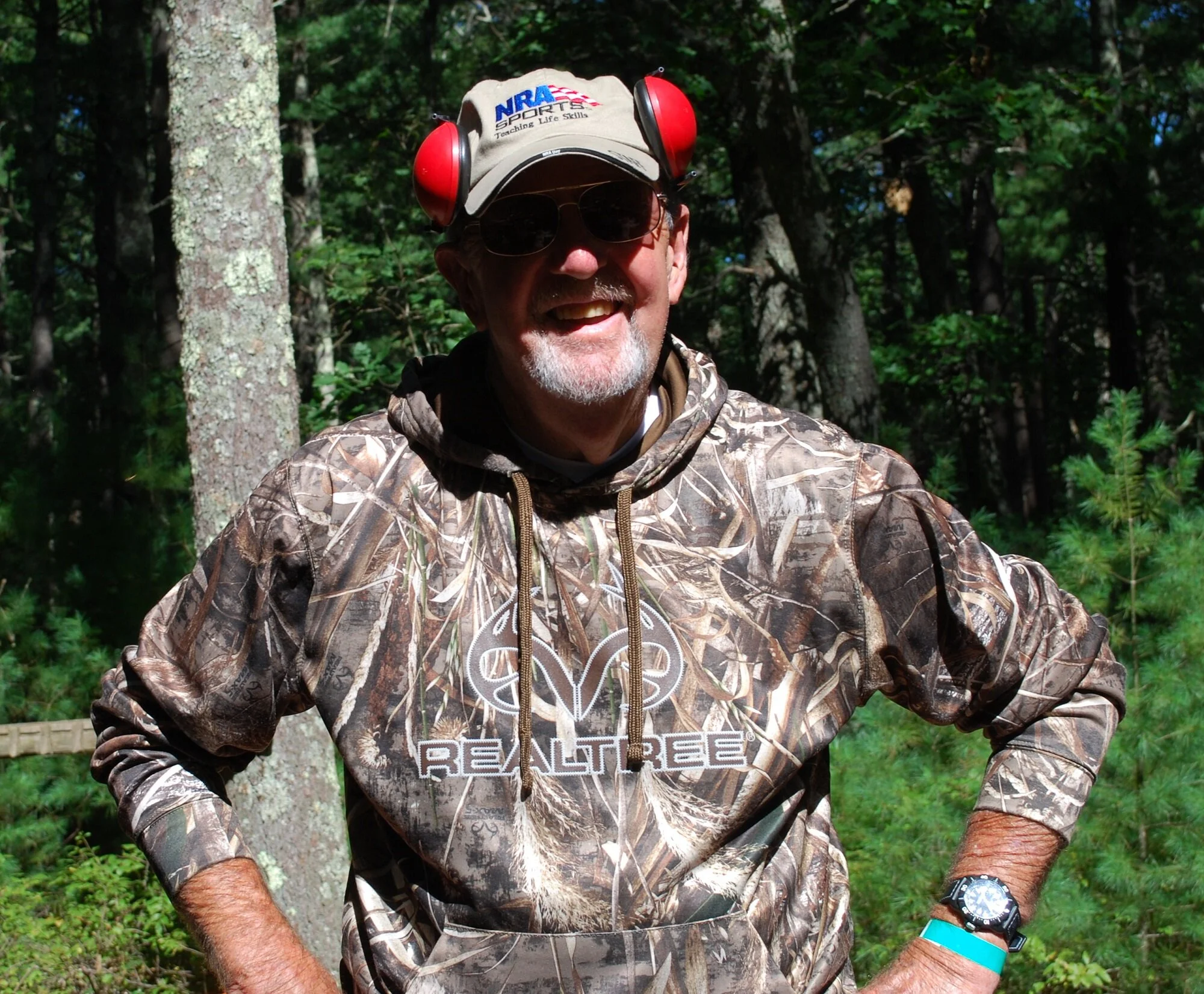 A smiling man in sunglasses and camouflage clothing standing outdoors among trees, wearing a beige cap with red ear protection, a watch, and a turquoise wristband.