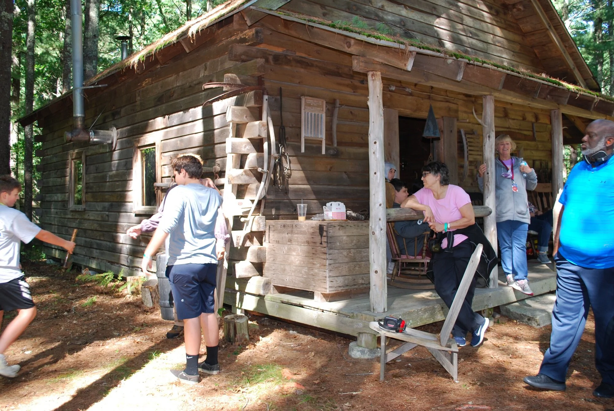 Group of people, including children and adults, gathered outside a rustic wooden cabin in a forest setting. Some are sitting on the porch, others are standing or walking nearby, engaging in conversation and activities.