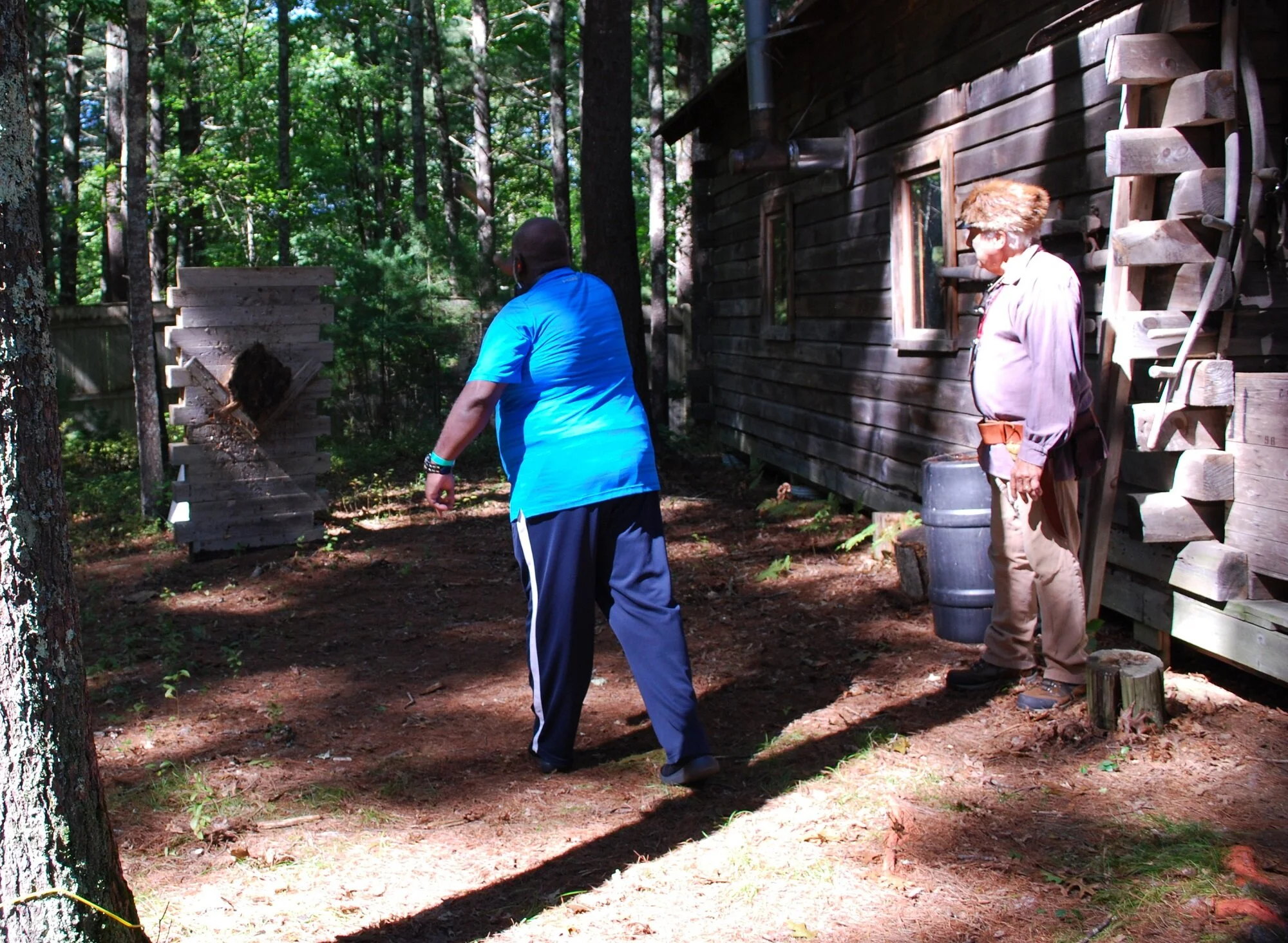 Two men stand outdoors near a wooden building in a wooded area, with one man wearing a blue shirt and black pants facing away and the other wearing a beige shirt and pants with sunglasses standing by the building.