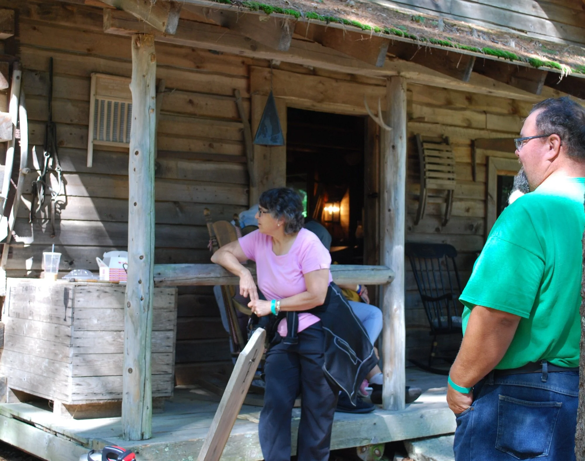 People standing on a wooden porch of a rustic cabin with tools hanging on the wall and a window with a deer antler above it.