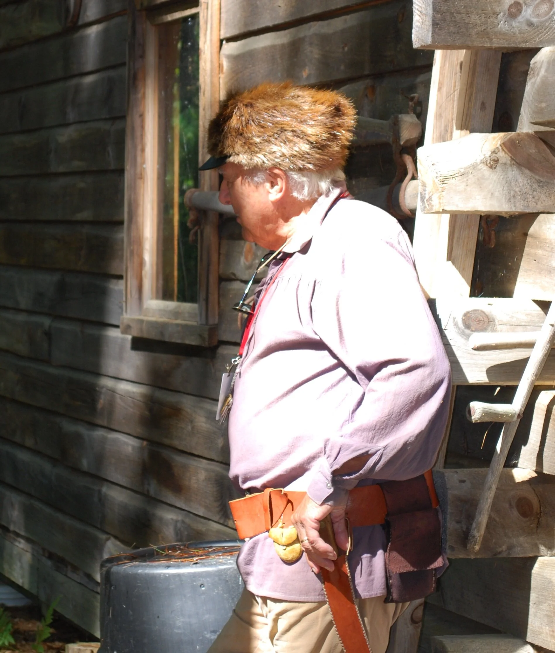 A man wearing a fur hat, light purple shirt, and tan pants, standing outside a wooden cabin, holding a leather belt and glasses, with a window and ladders visible on the cabin wall.