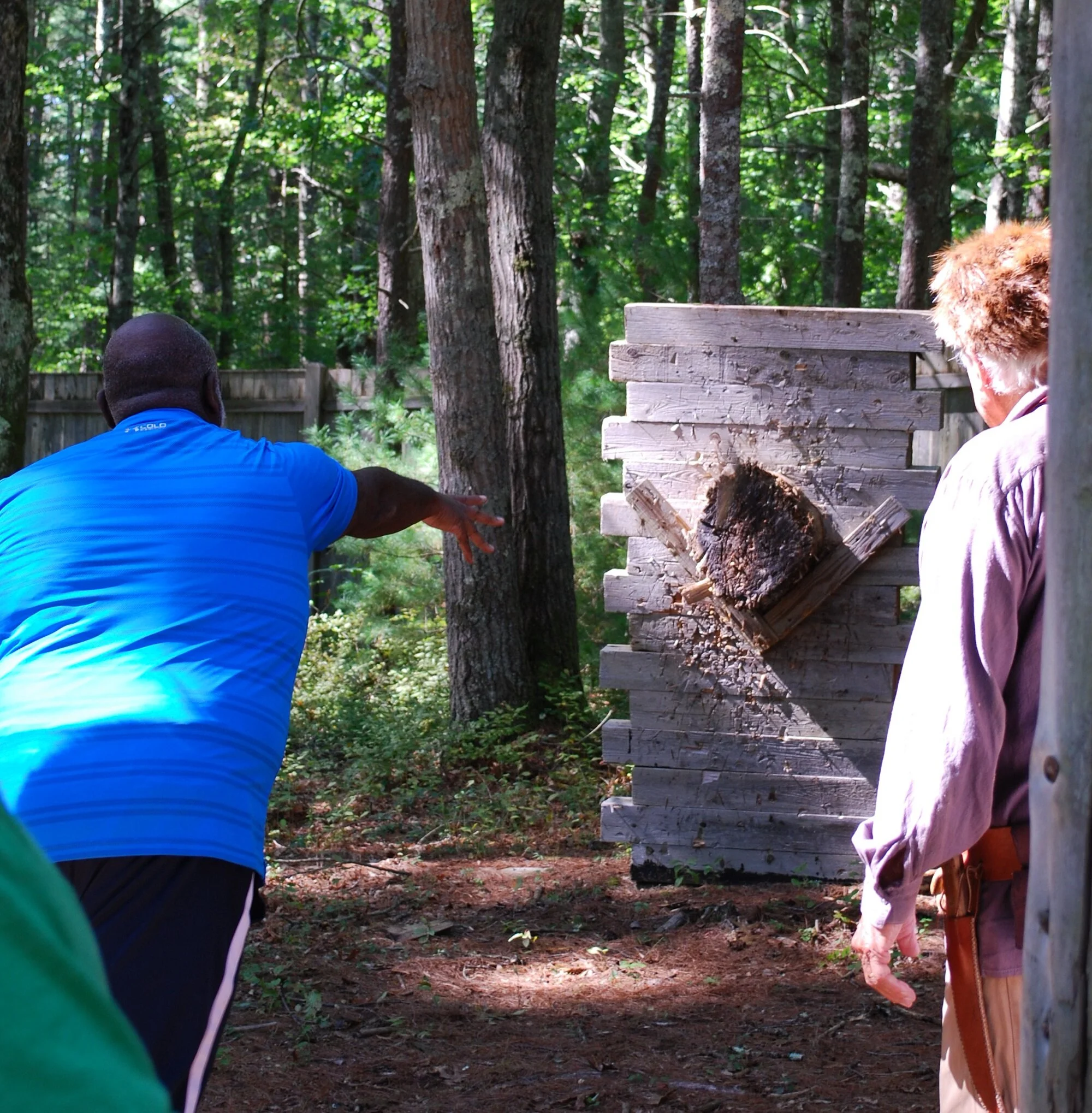 Two men participating in an axe-throwing activity outdoors among trees. One man is mid-throw with his arm extended, while the other observes. There is a wooden target with a log placed on it, set up in a forested area.
