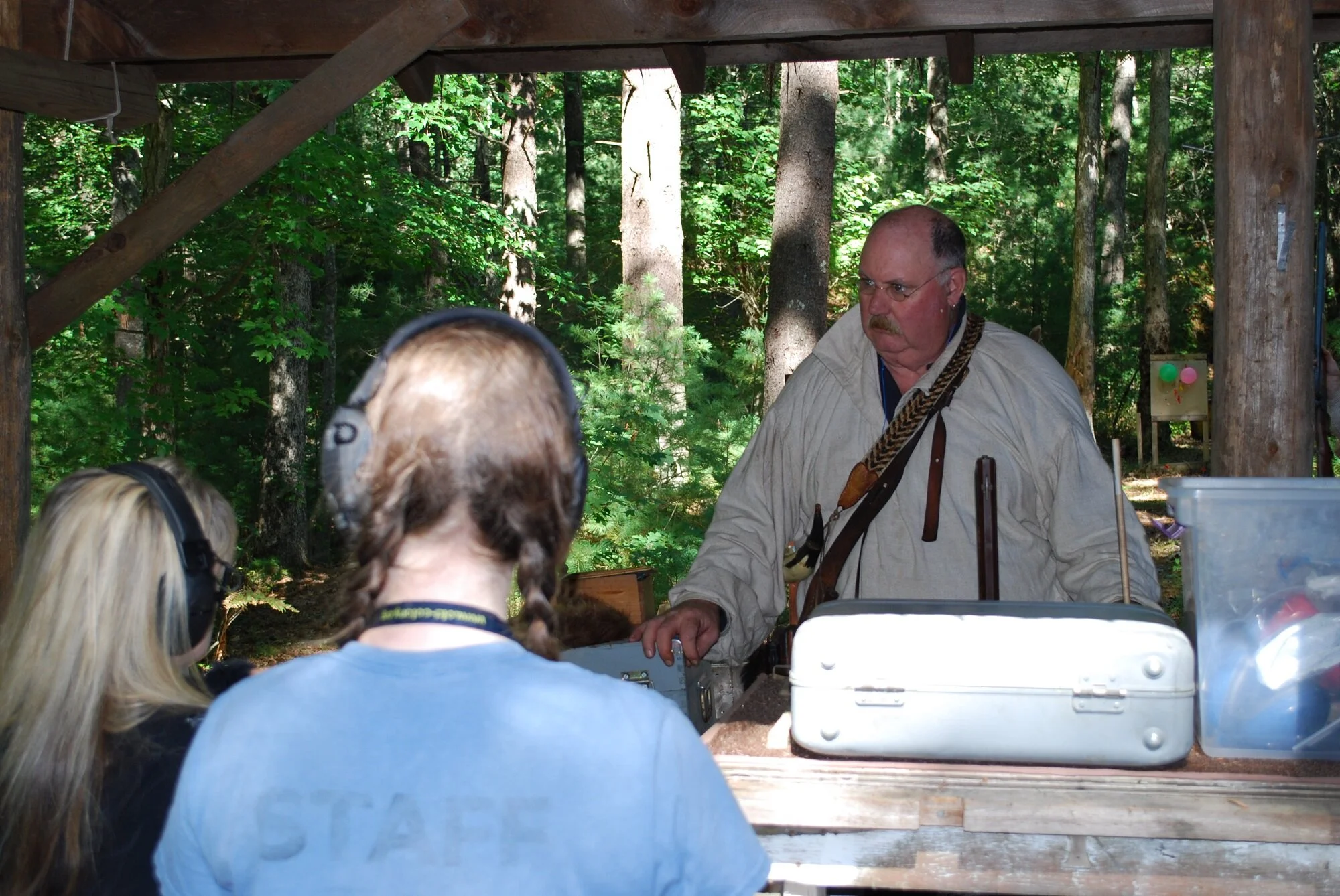 Two children with headphones learning from an older man in an outdoor wooded setting under a wooden shelter.