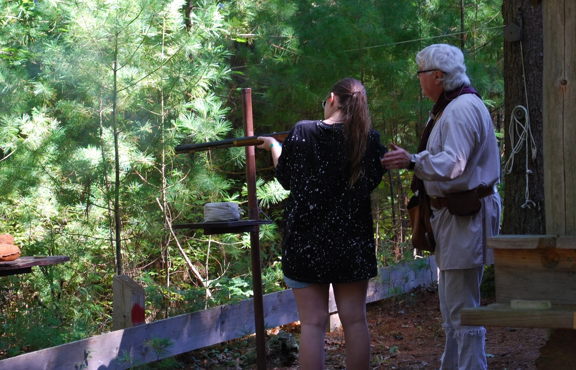 A young woman and an older man outdoors in a wooded area, with the woman aiming a long gun at a target.