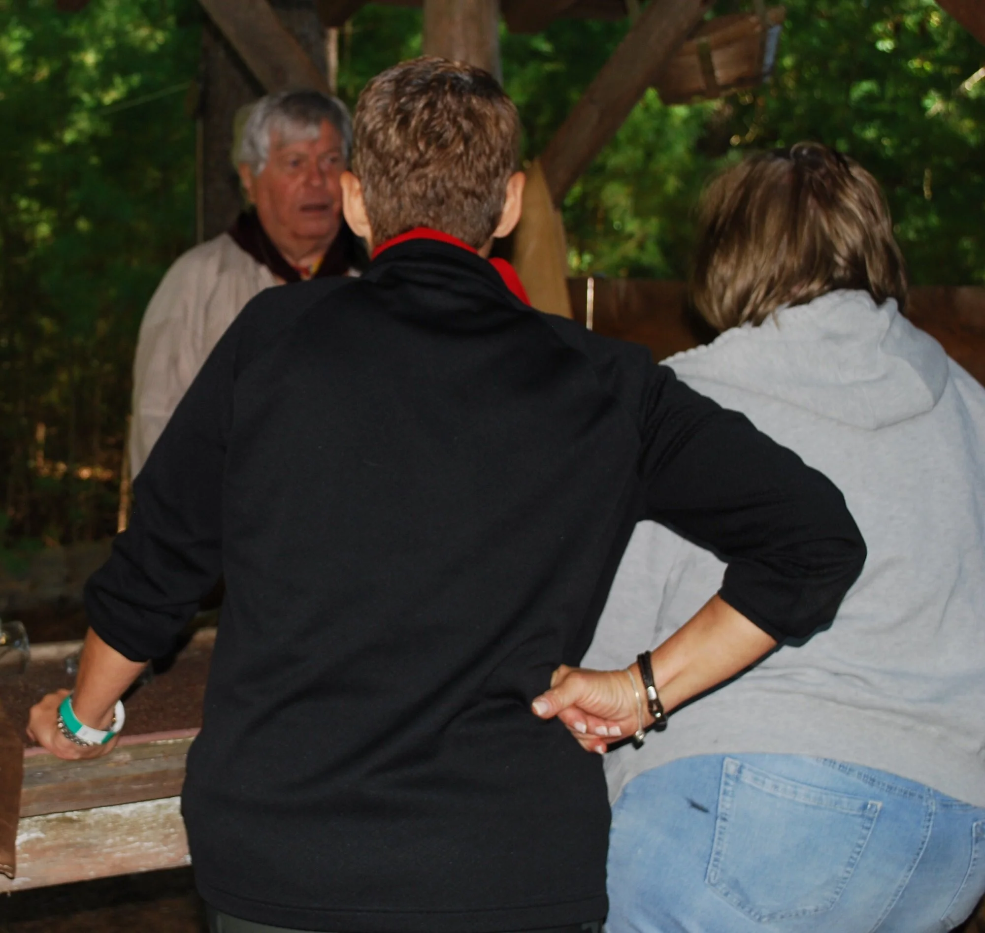 Three people are engaged in conversation outdoors near trees and wooden structures.