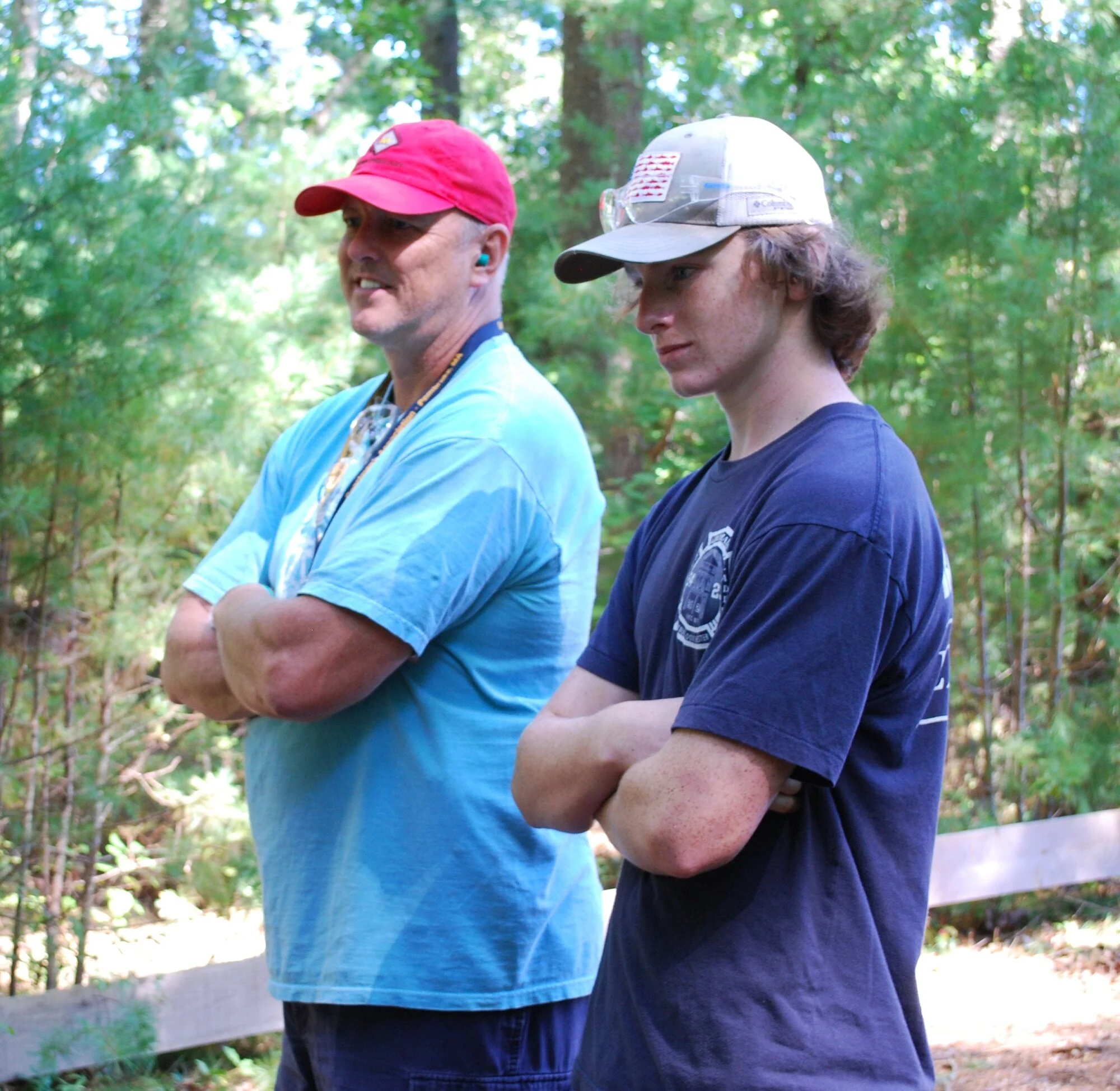 Two men standing outdoors in a wooded area, wearing casual t-shirts and hats with crossed arms.