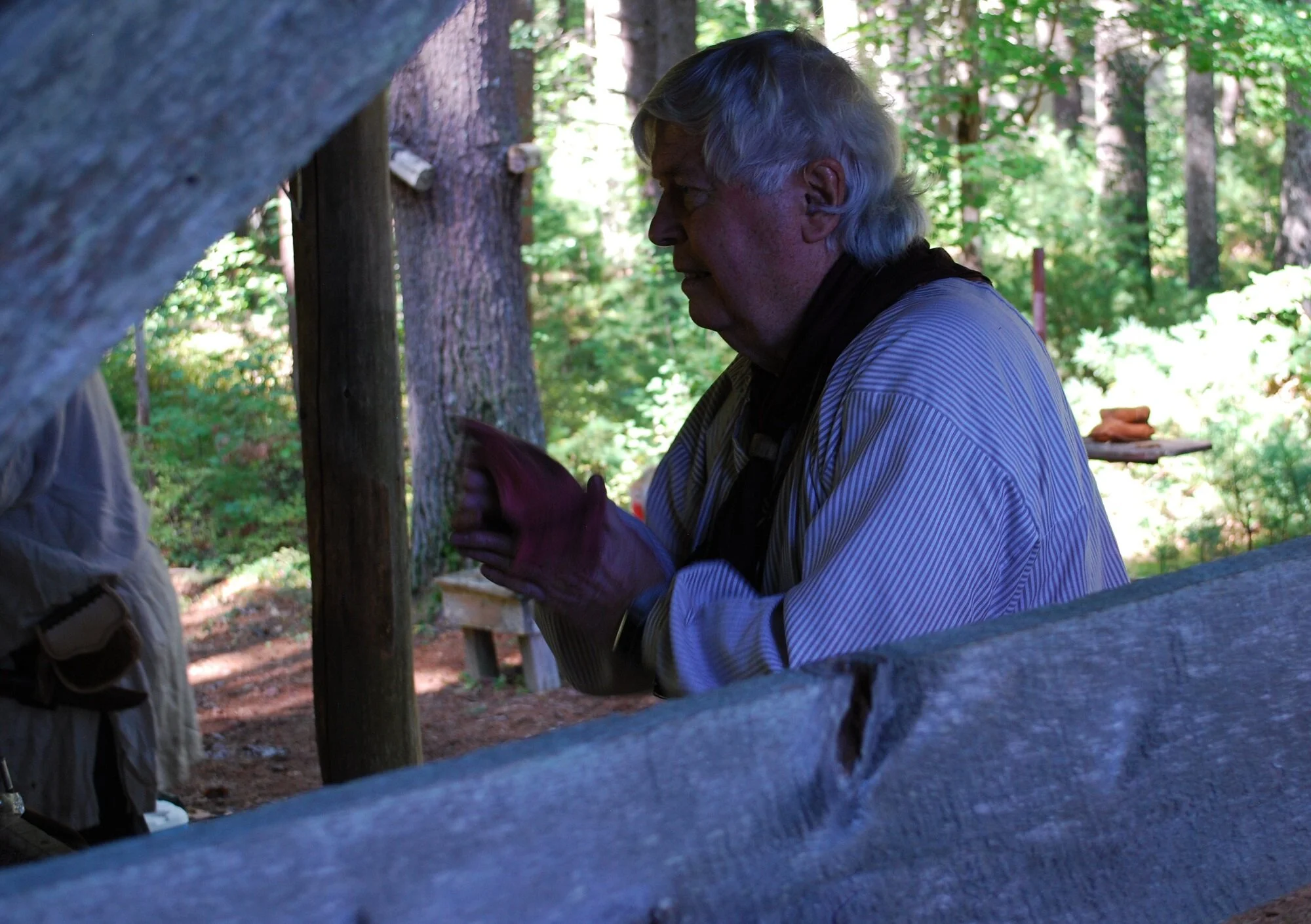 An elderly man with white hair and a striped shirt, wearing red gloves, is standing outdoors among trees and greenery, looking down and holding an object.