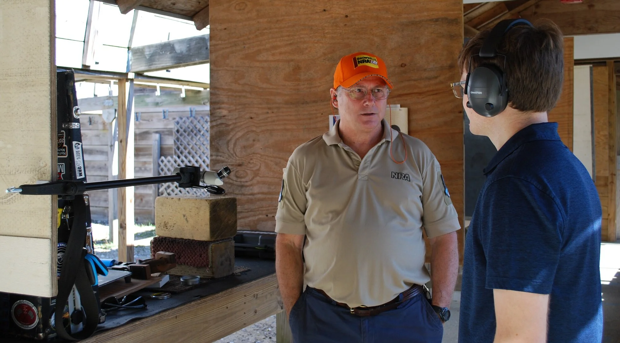 Two men having a conversation at an outdoor shooting range. One man wears a beige NRA shirt and orange NRA day hat, the other man wears a blue shirt and large hearing protection. Shooting equipment and targets are visible in the background.