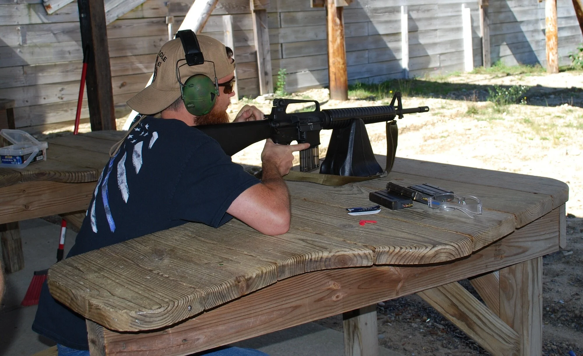 Man aiming a rifle at a shooting range, wearing headphones, a cap, and sunglasses, sitting at a wooden table with ammunition and safety glasses.