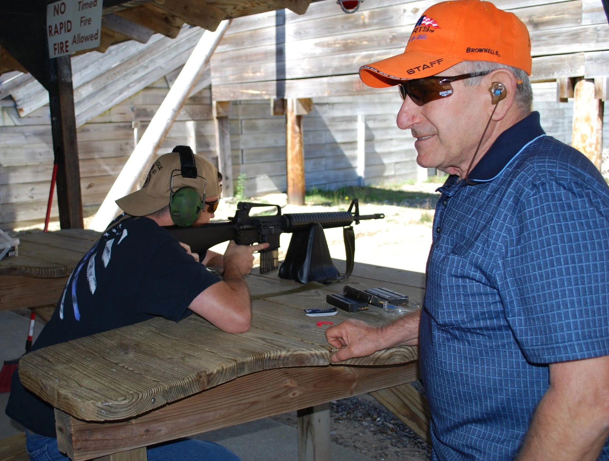 A man in a blue shirt and orange cap is facing a shooting range, observing a young man aiming a rifle on a wooden table at an outdoor shooting range with wooden fencing.