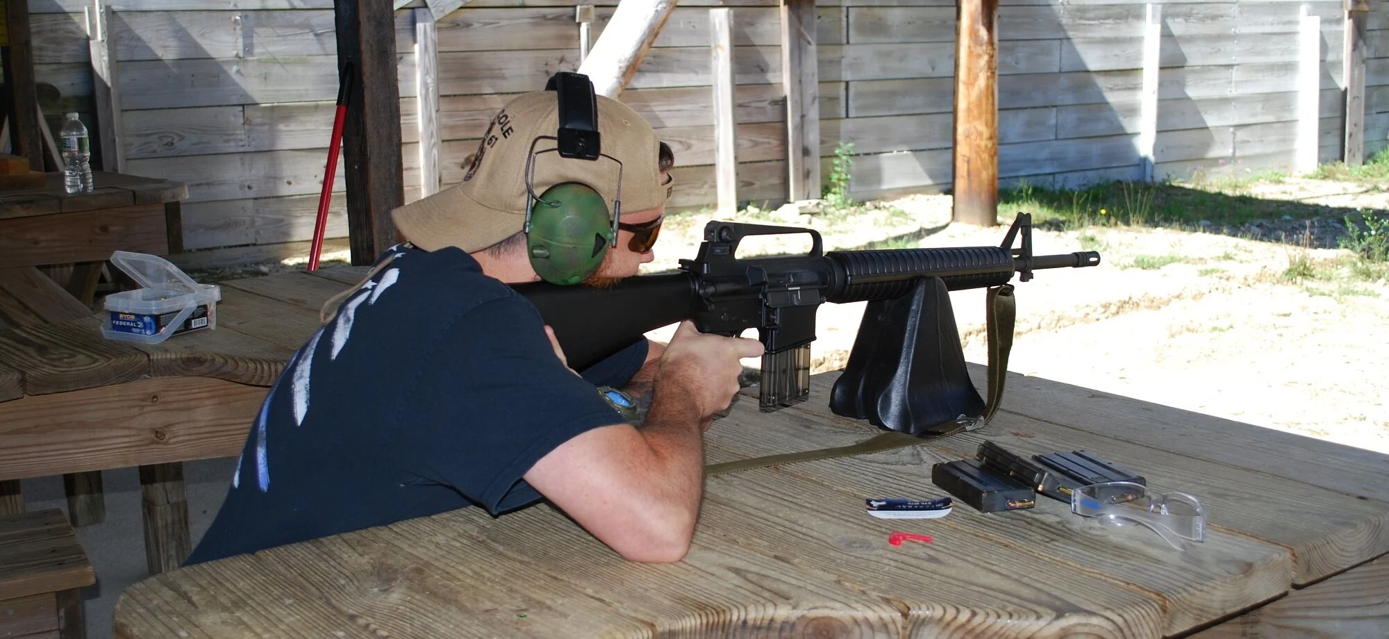 A man is aiming a rifle at a shooting range, lying on a wooden table, wearing protective ear and eye gear, with shotgun shells and a magazine nearby.